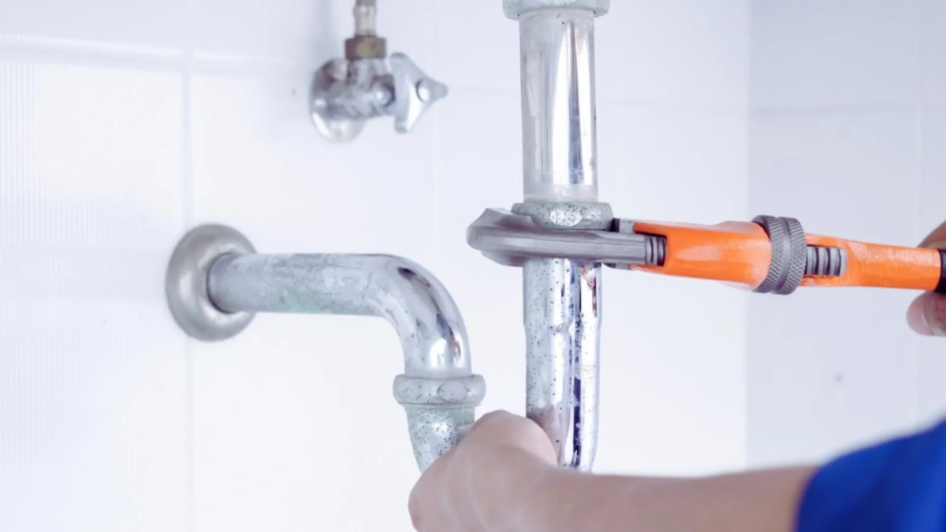 Plumber tightening a pipe under a sink using an orange pipe wrench and hands visible.