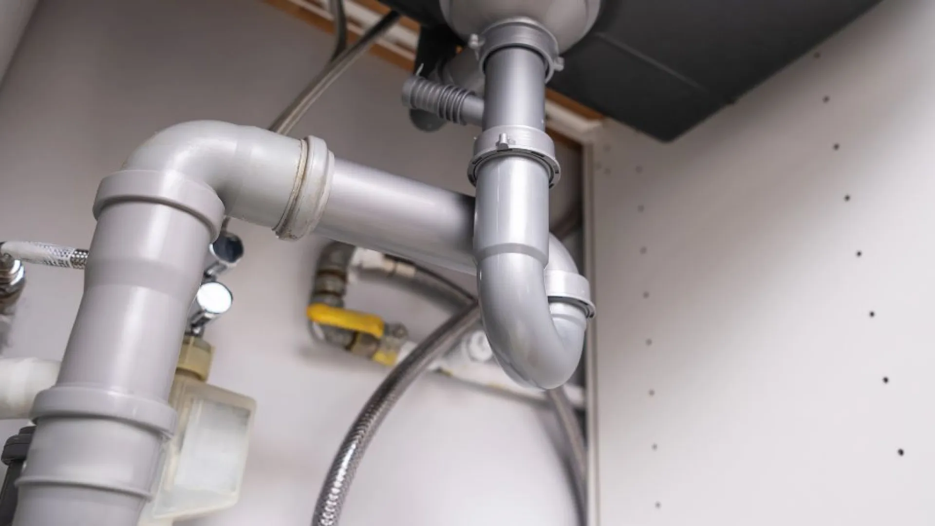 Close-up of gray under-sink plumbing pipes and fittings with flexible water hoses in a cabinet space.