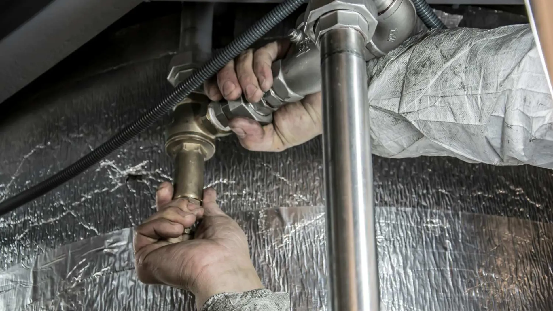 Technician repairing or installing plumbing pipes under a sink with metallic insulation in the background