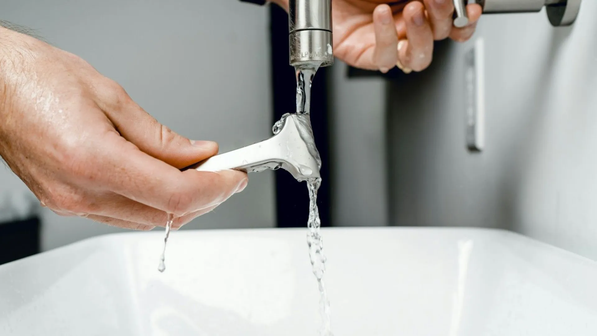 Person rinsing a metal safety razor under running water from a bathroom faucet above a white sink.