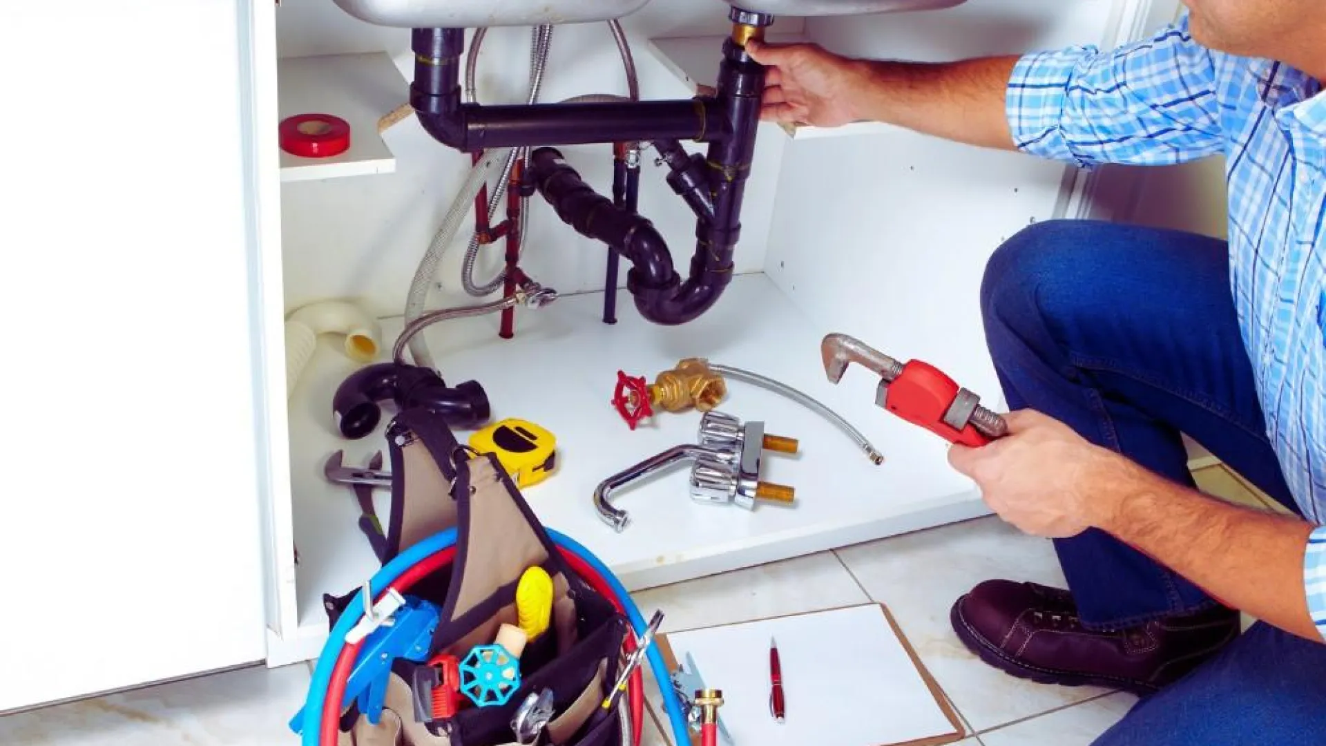 Plumber repairing kitchen sink pipes with tool bag and plumbing parts on tiled floor under sink