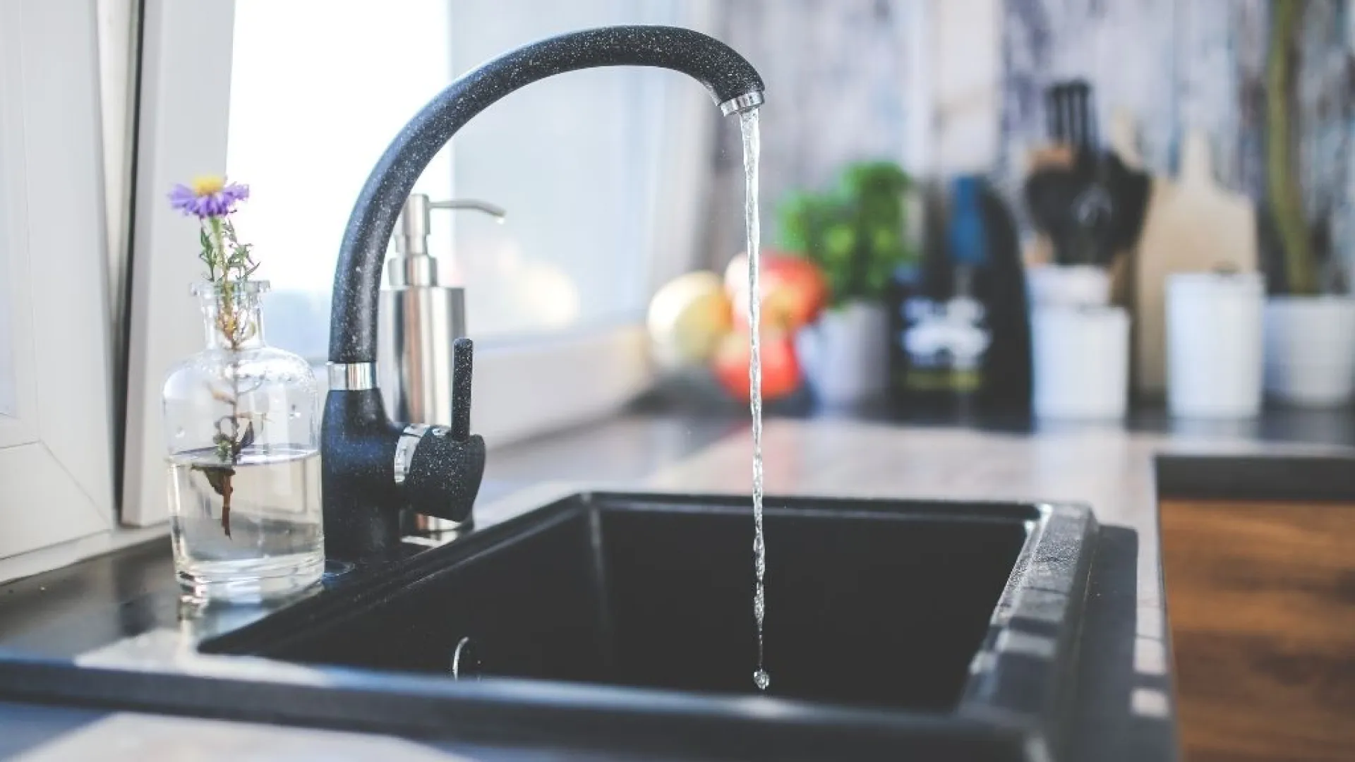 Modern black kitchen faucet with running water over a black sink with blurred kitchen background and vase