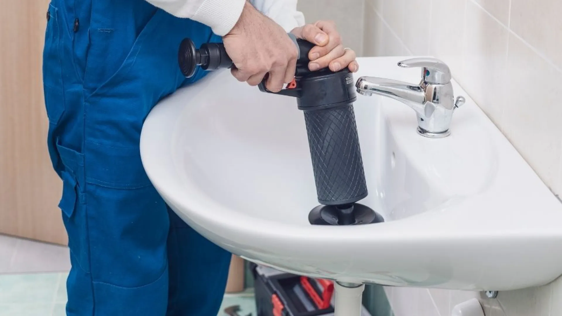 Plumber in blue overalls using a manual drain auger to unclog a white bathroom sink drain effectively.