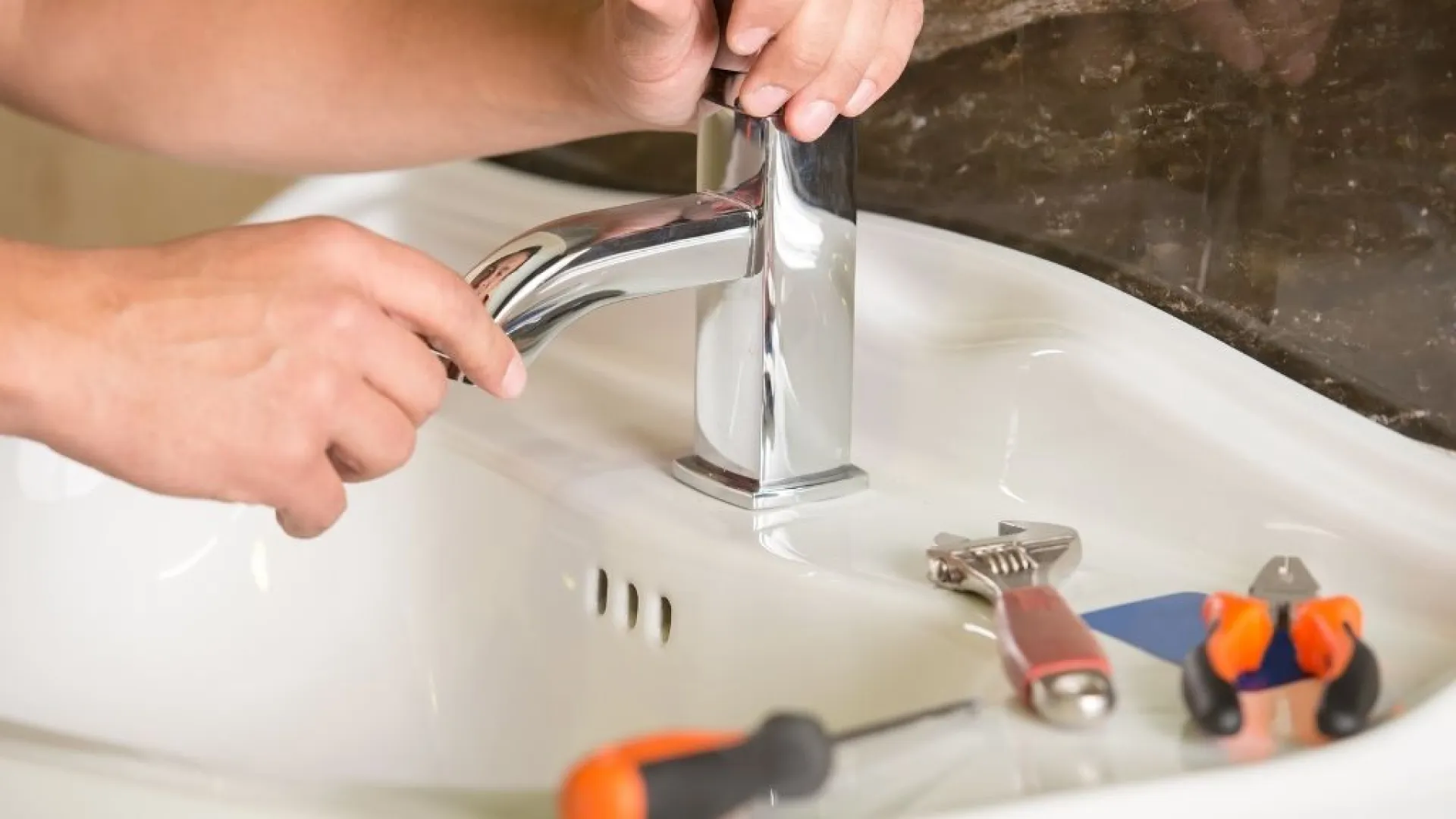 Hands repairing a modern chrome faucet on a white sink with tools including a wrench and screwdriver nearby