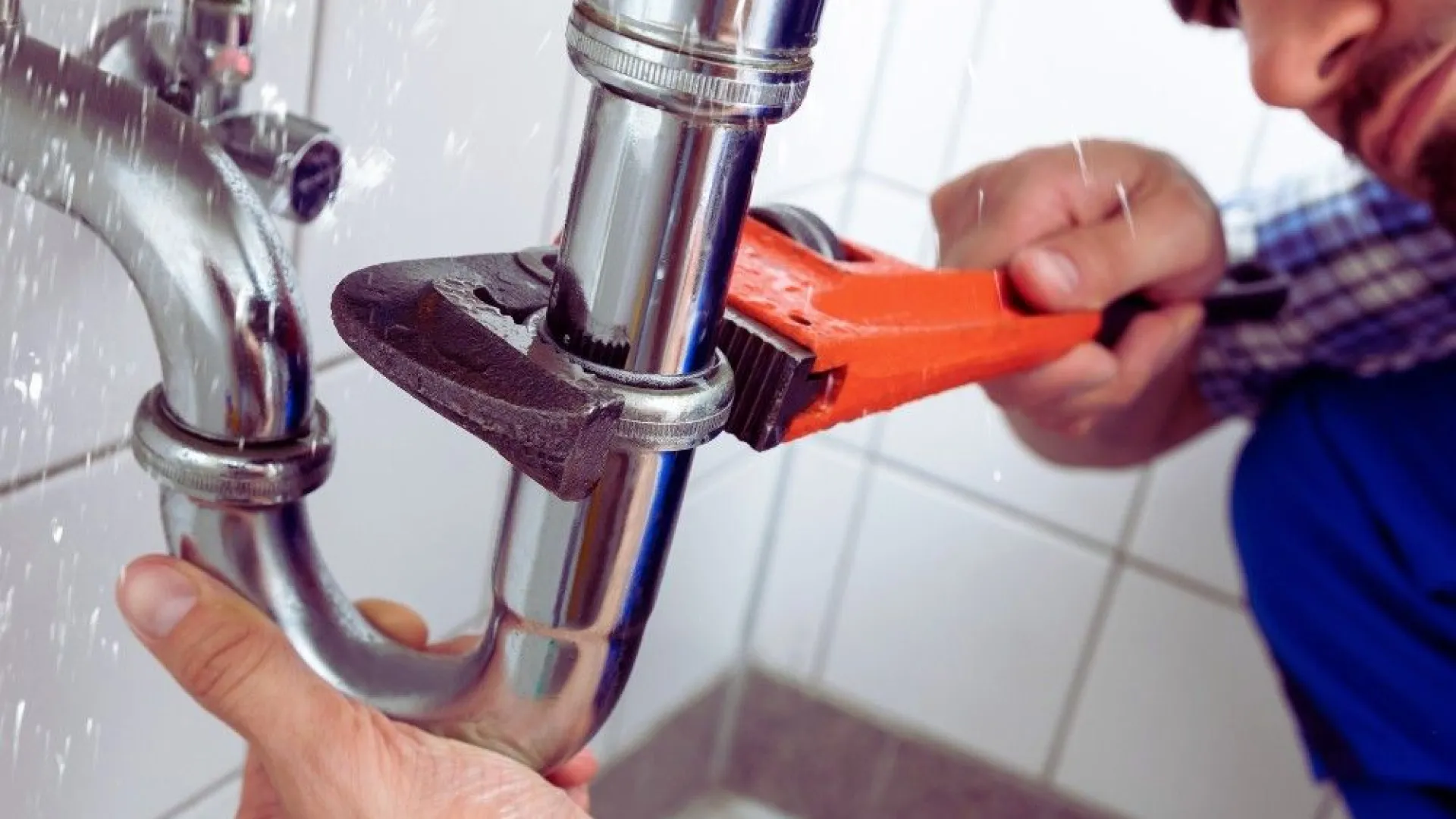 Plumber urgently fixing leaking bathroom pipe under sink with wrench and water spraying around.