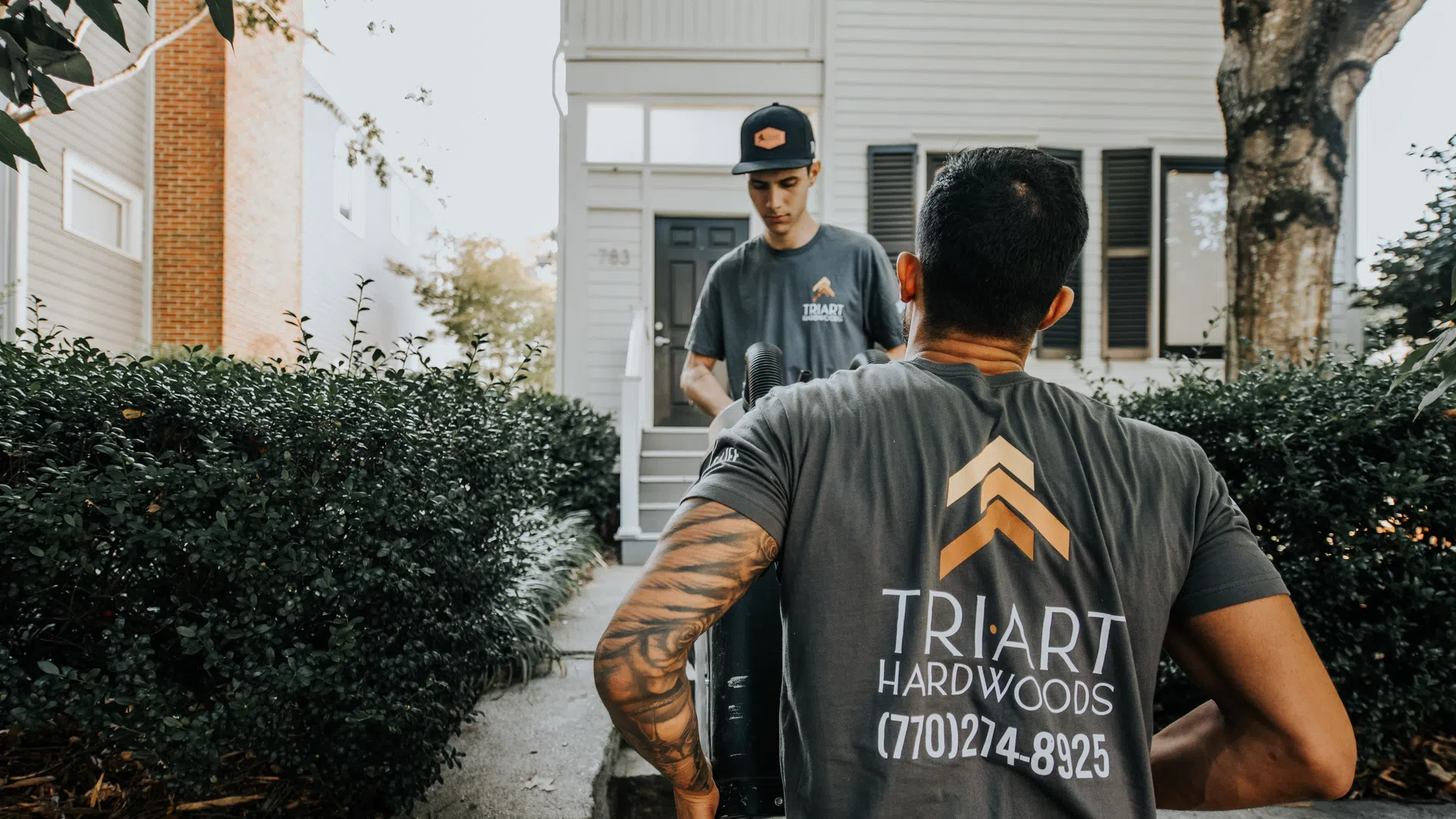 Two workers in Triart Hardwoods shirts moving equipment outside a house with bushes and stone wall.