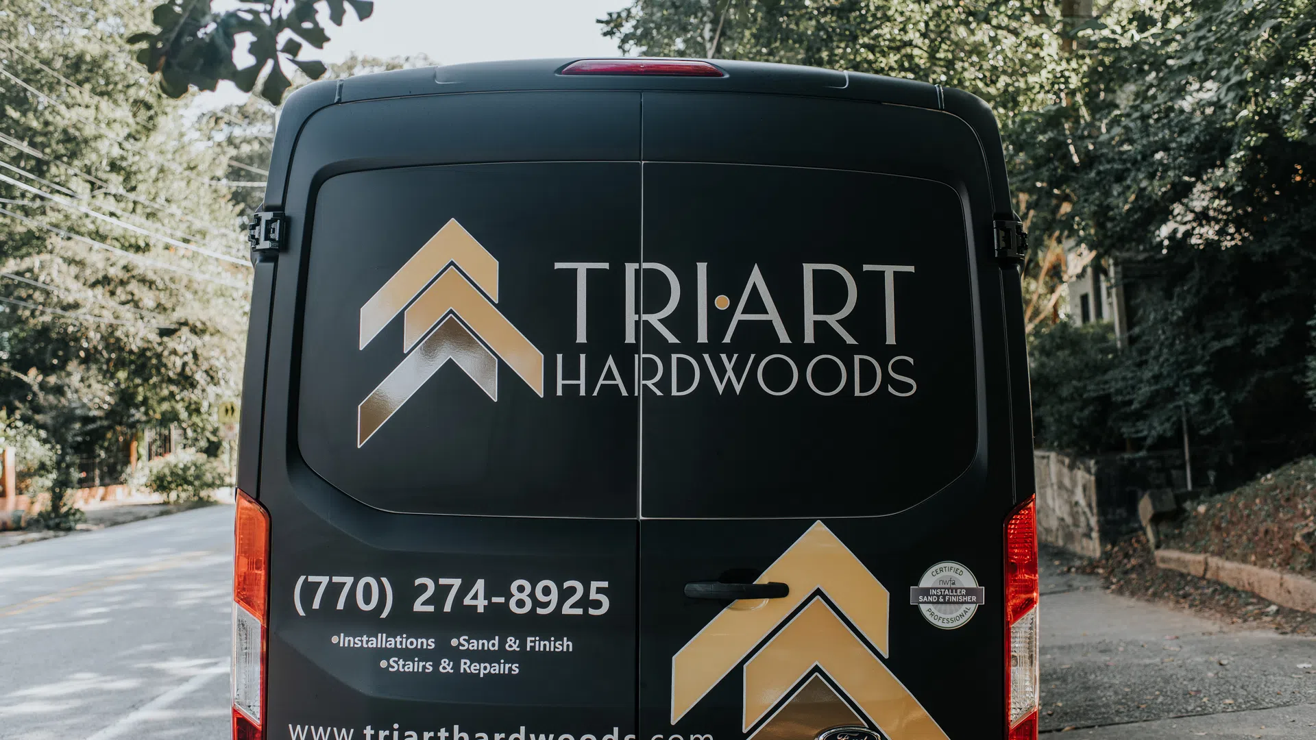 Black van with Tri Art Hardwoods logo, contact number, services, and website displayed on back doors.