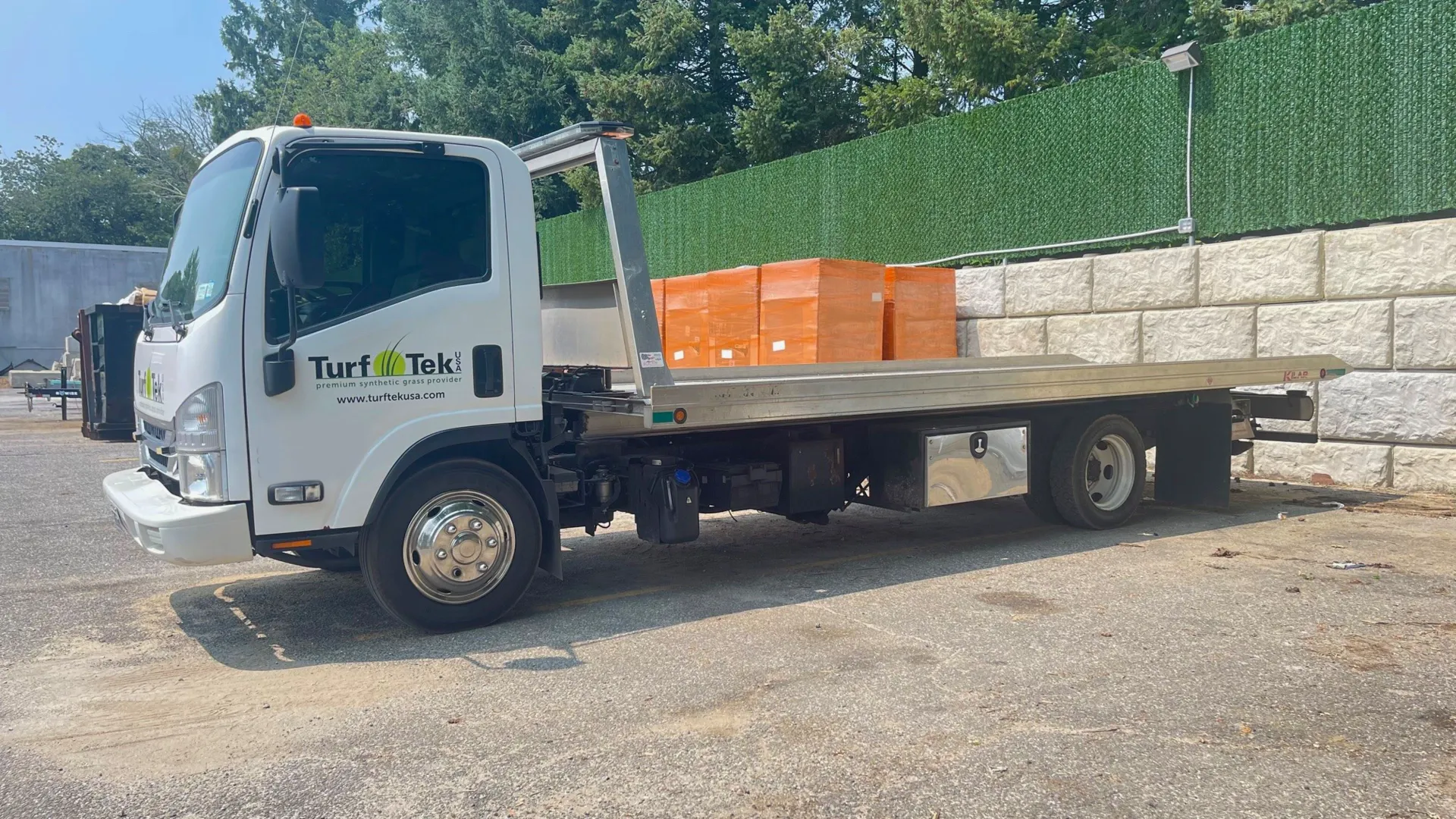 White flatbed tow truck with Turf Tek logo parked next to stone wall and hedge under clear sky.