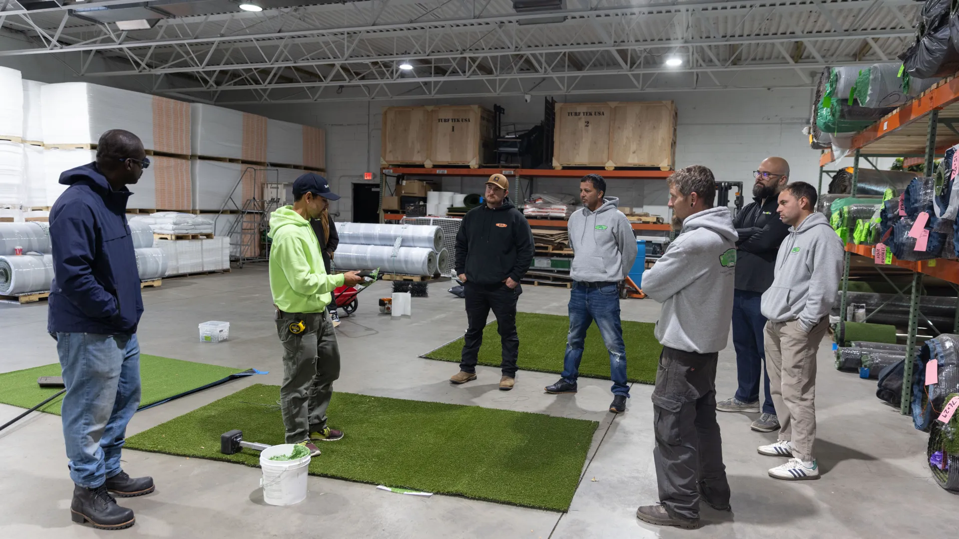 Group of workers receiving instructions inside a warehouse with artificial turf samples and supplies.