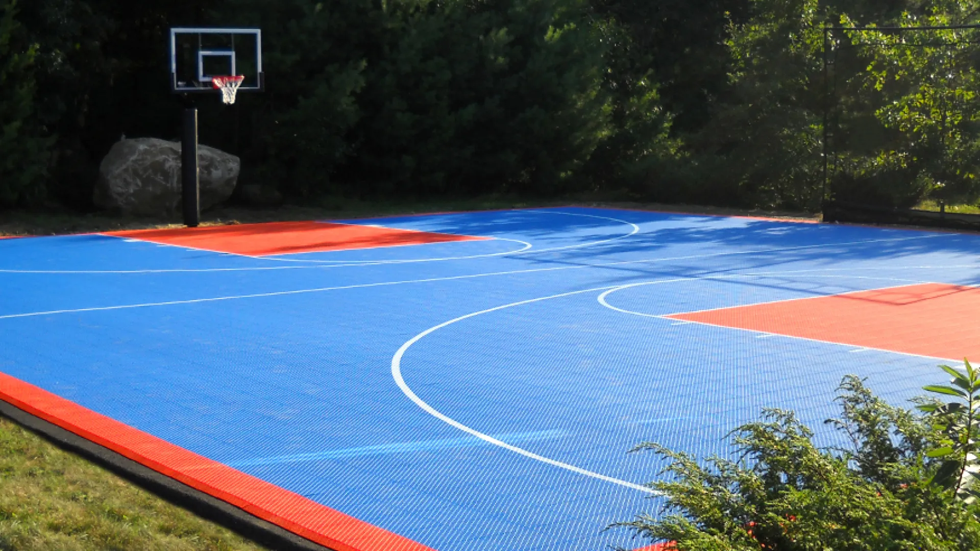 Outdoor basketball court with blue and orange flooring surrounded by trees and greenery on a sunny day