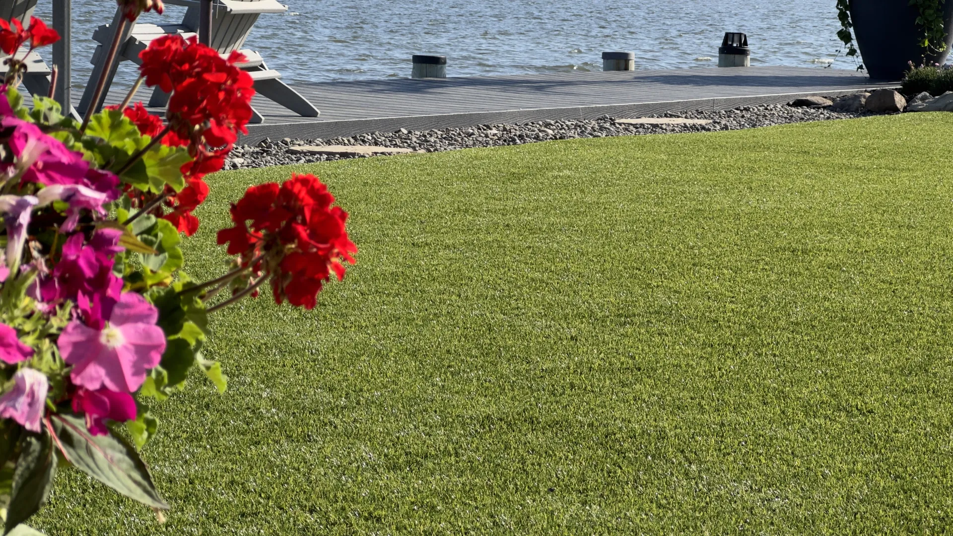 Green lawn with bright red and pink flowers near a wooden dock by calm water on a sunny day