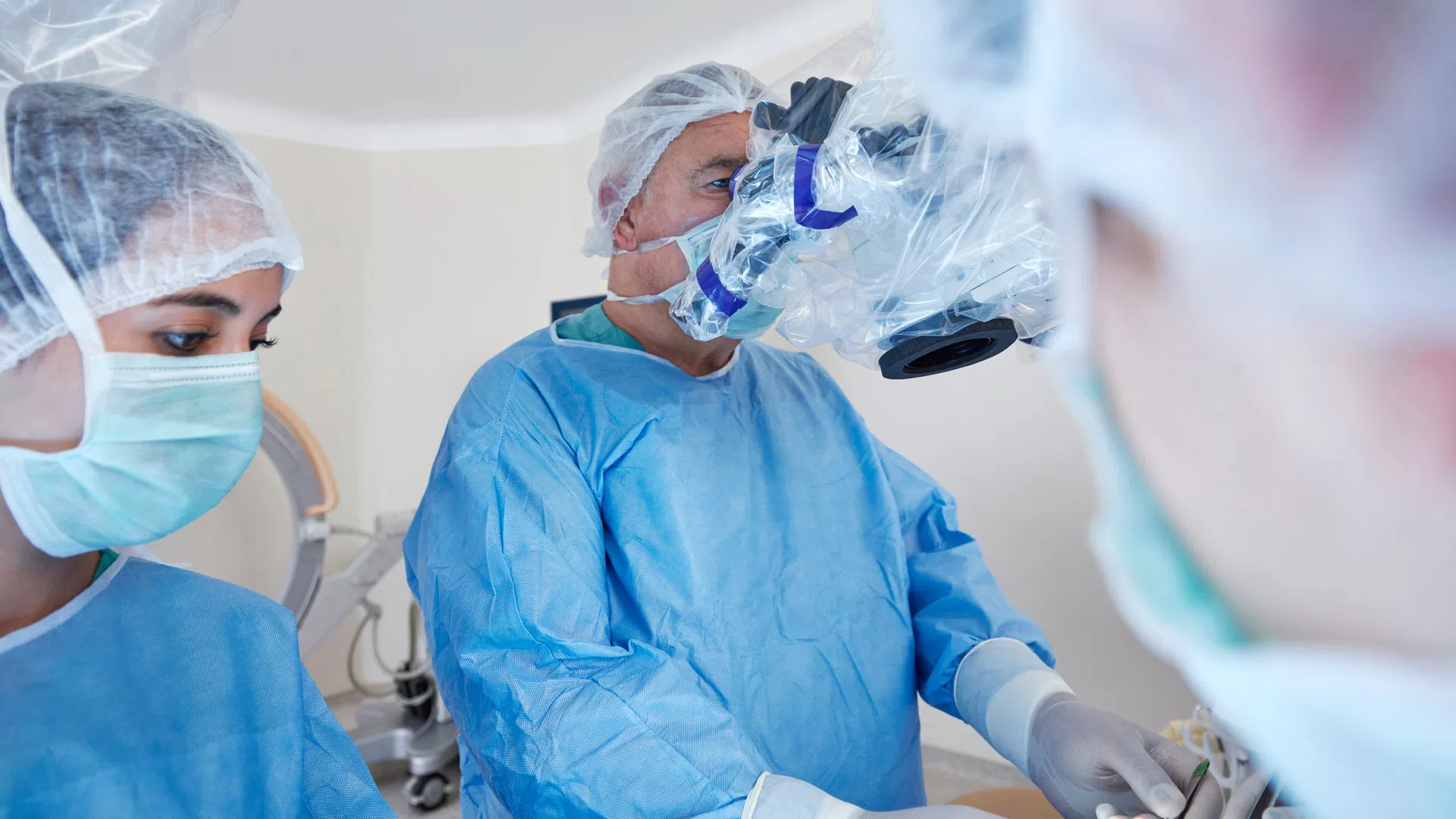 Surgeons in blue scrubs and masks perform a medical procedure using a surgical microscope in an operating room.