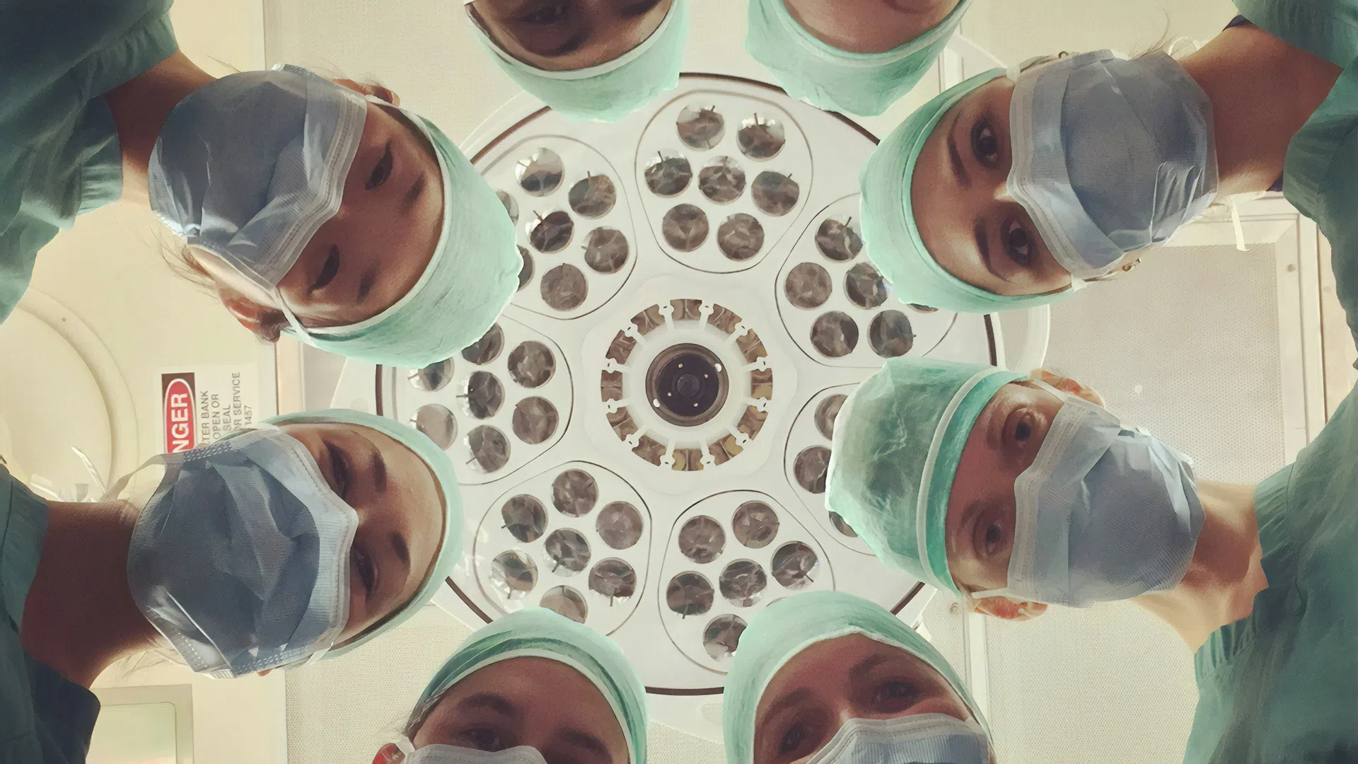 Surgeons wearing masks and scrubs looking down under circular operating room lights in a hospital.