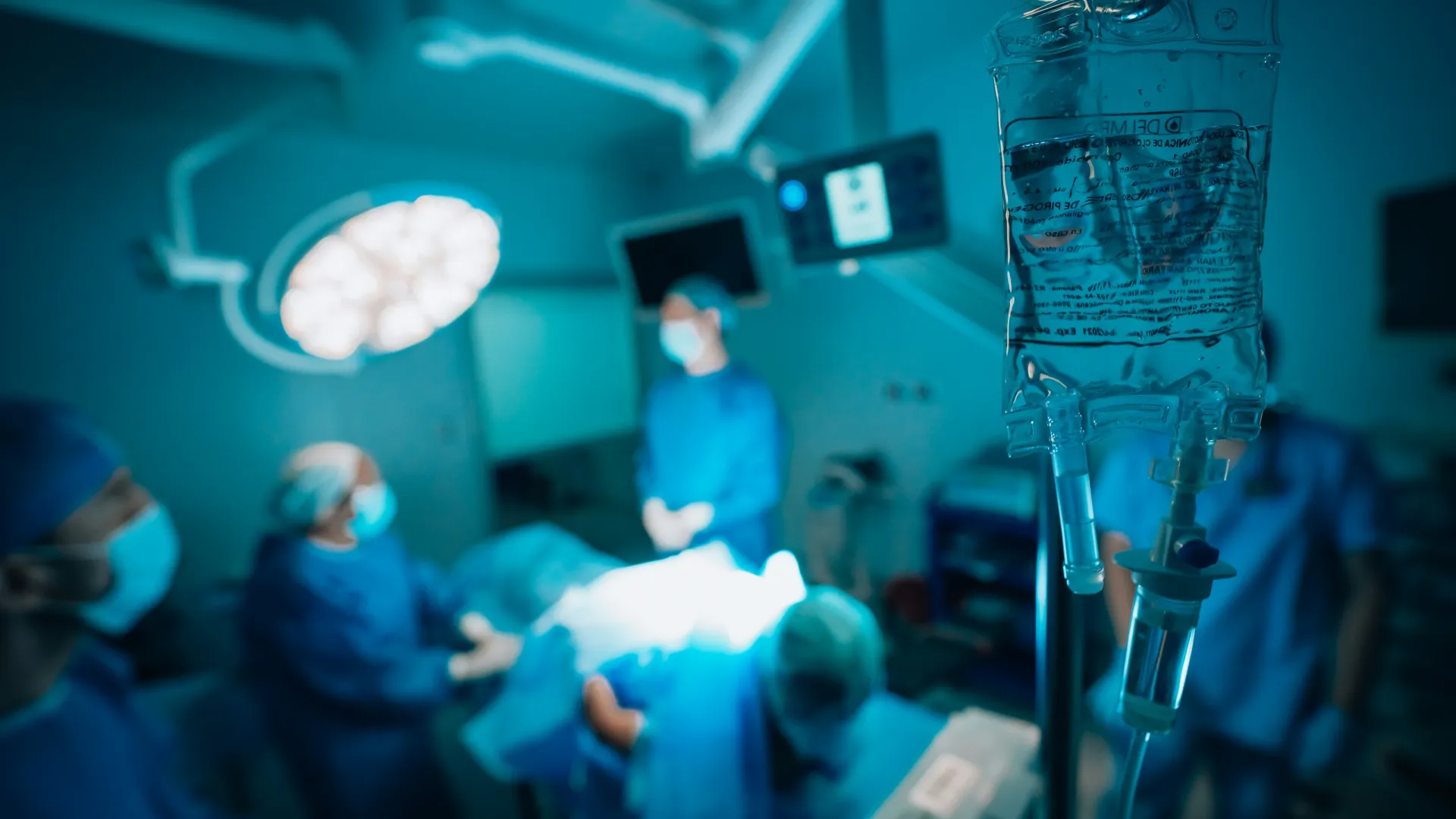Intravenous drip in focus with blurred surgical team performing operation in sterile operating room.