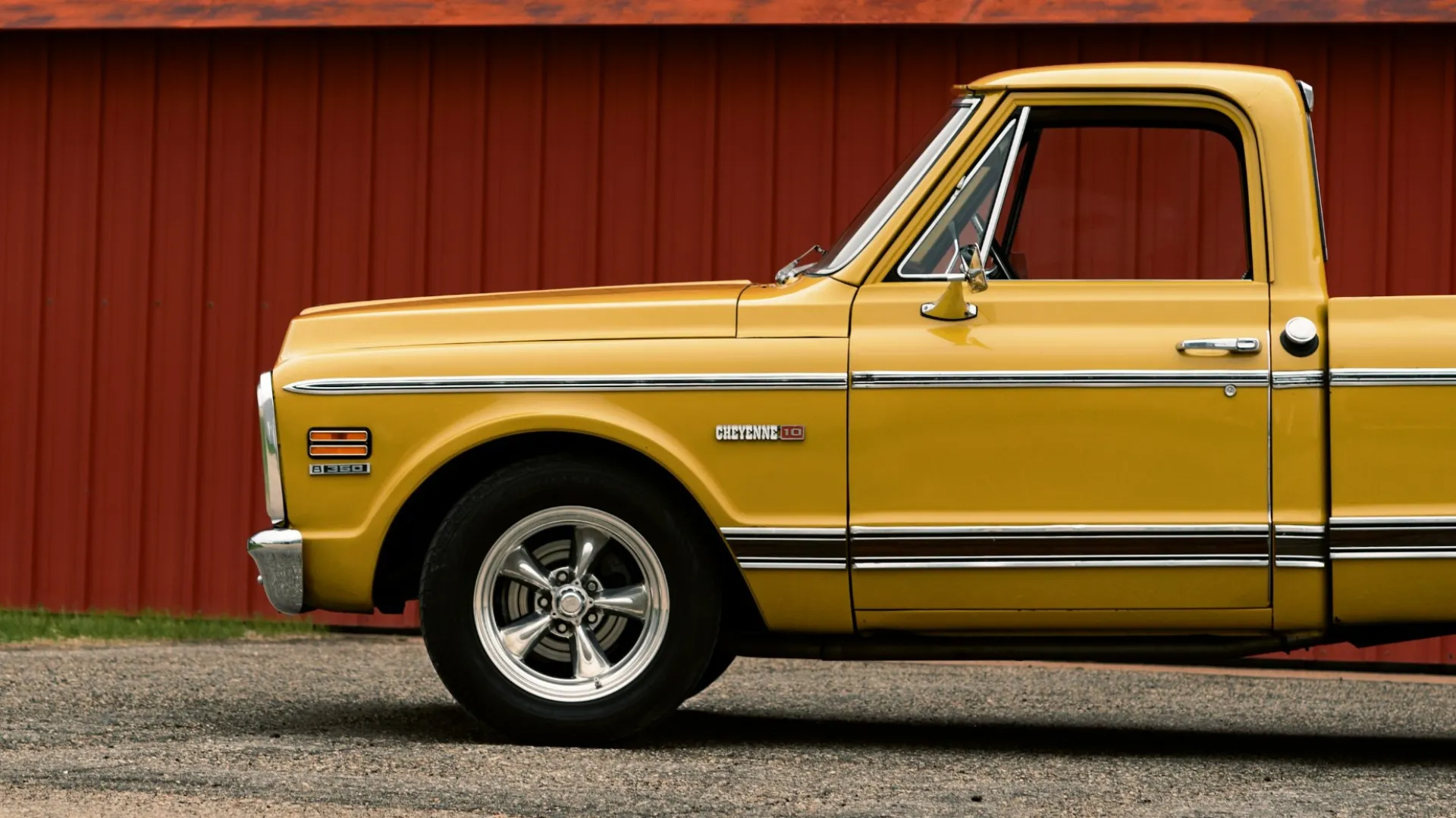 Side view of a vintage yellow Chevrolet C10 pickup truck parked in front of a red metal building.