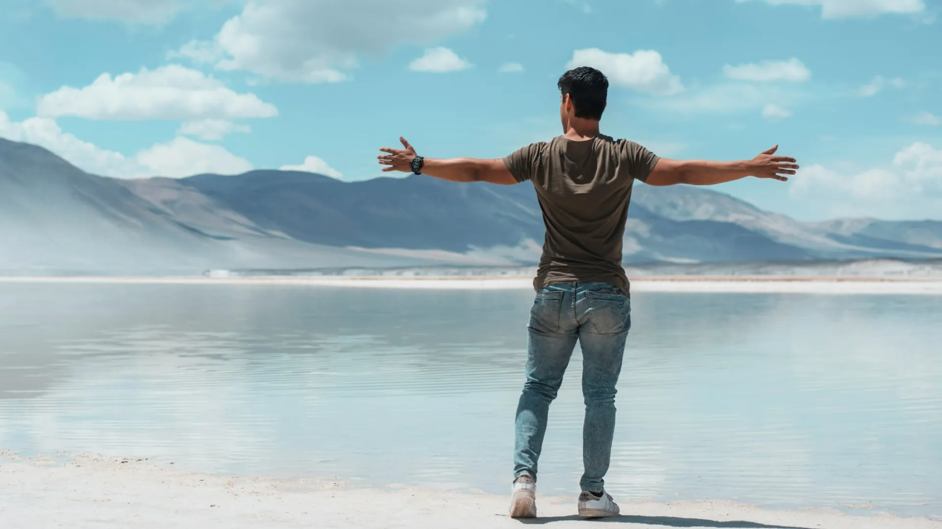 Man standing with arms outstretched by a calm lake with mountains and cloudy sky in the background.
