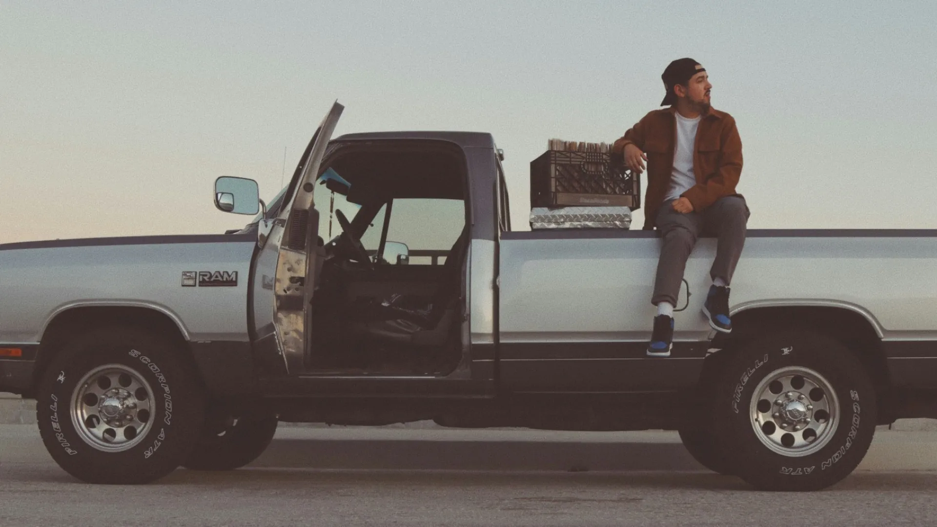 Man sitting on the bed of a silver pickup truck with the driver's door open during sunset.