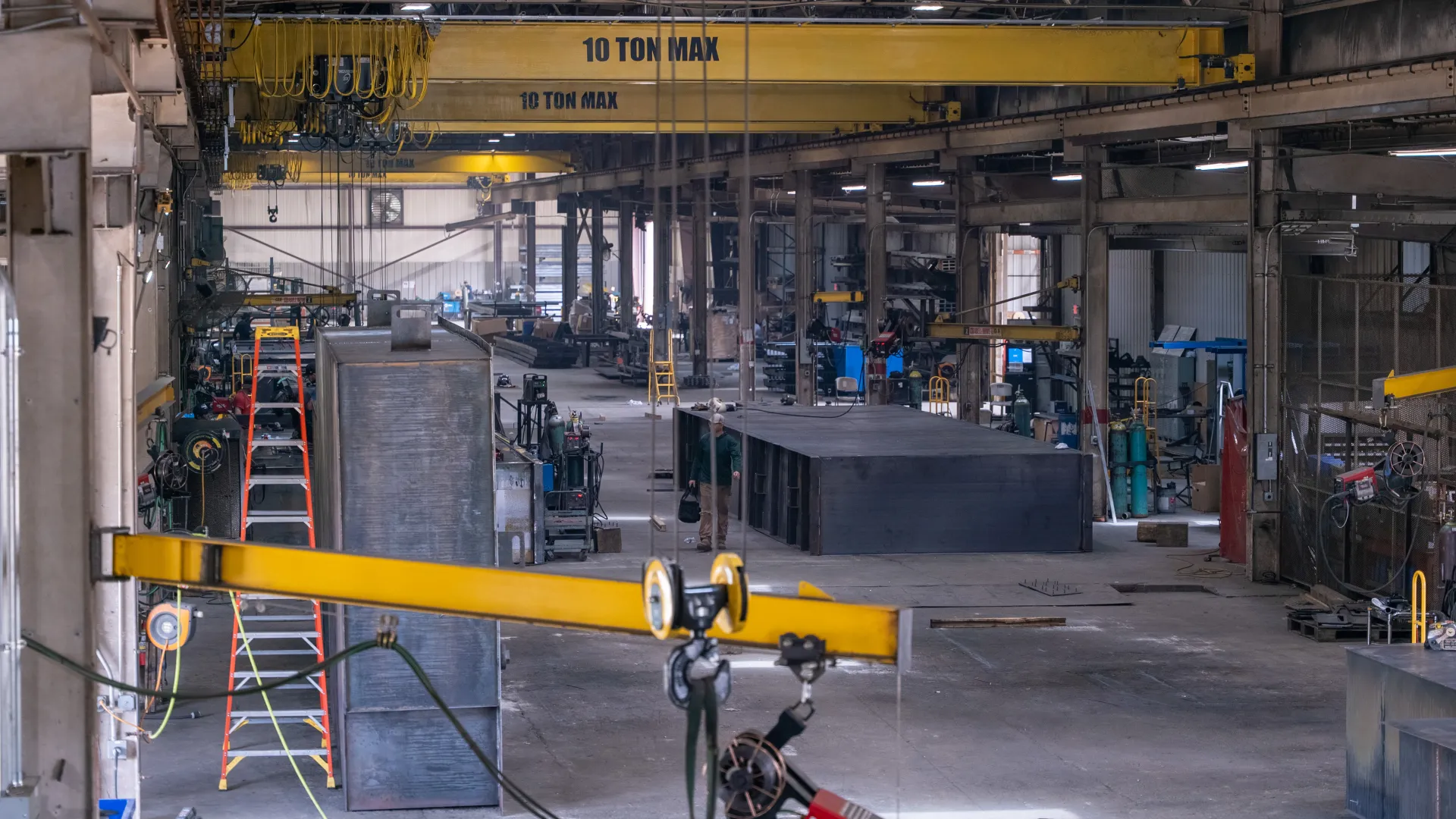 Industrial warehouse interior with overhead cranes, metal structures, ladders, and manufacturing equipment.
