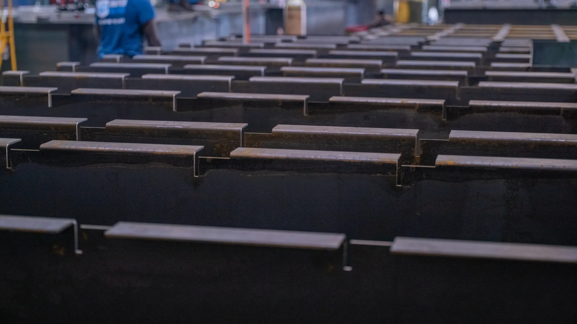 Rows of steel beams lined up in a factory with workers in the background assembling machinery.