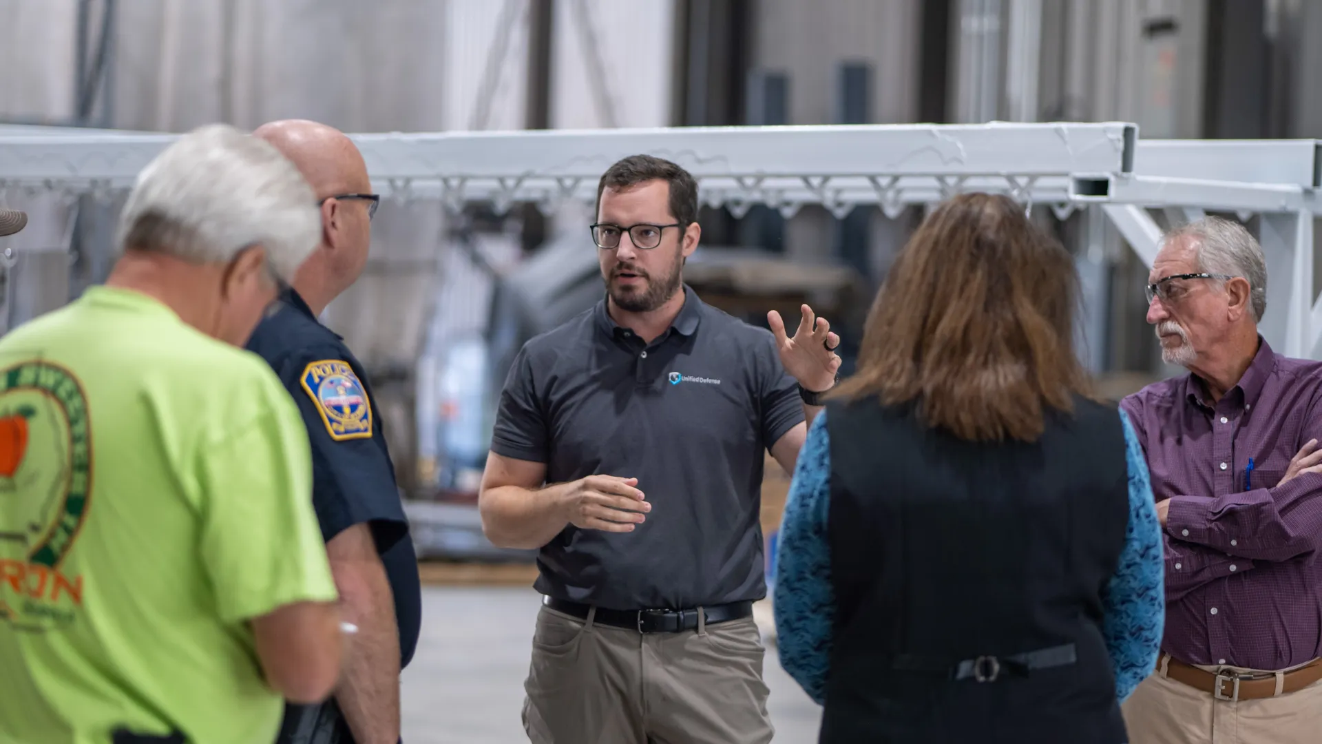 Man in black polo explaining to group of adults including police officer and elderly people in industrial setting.
