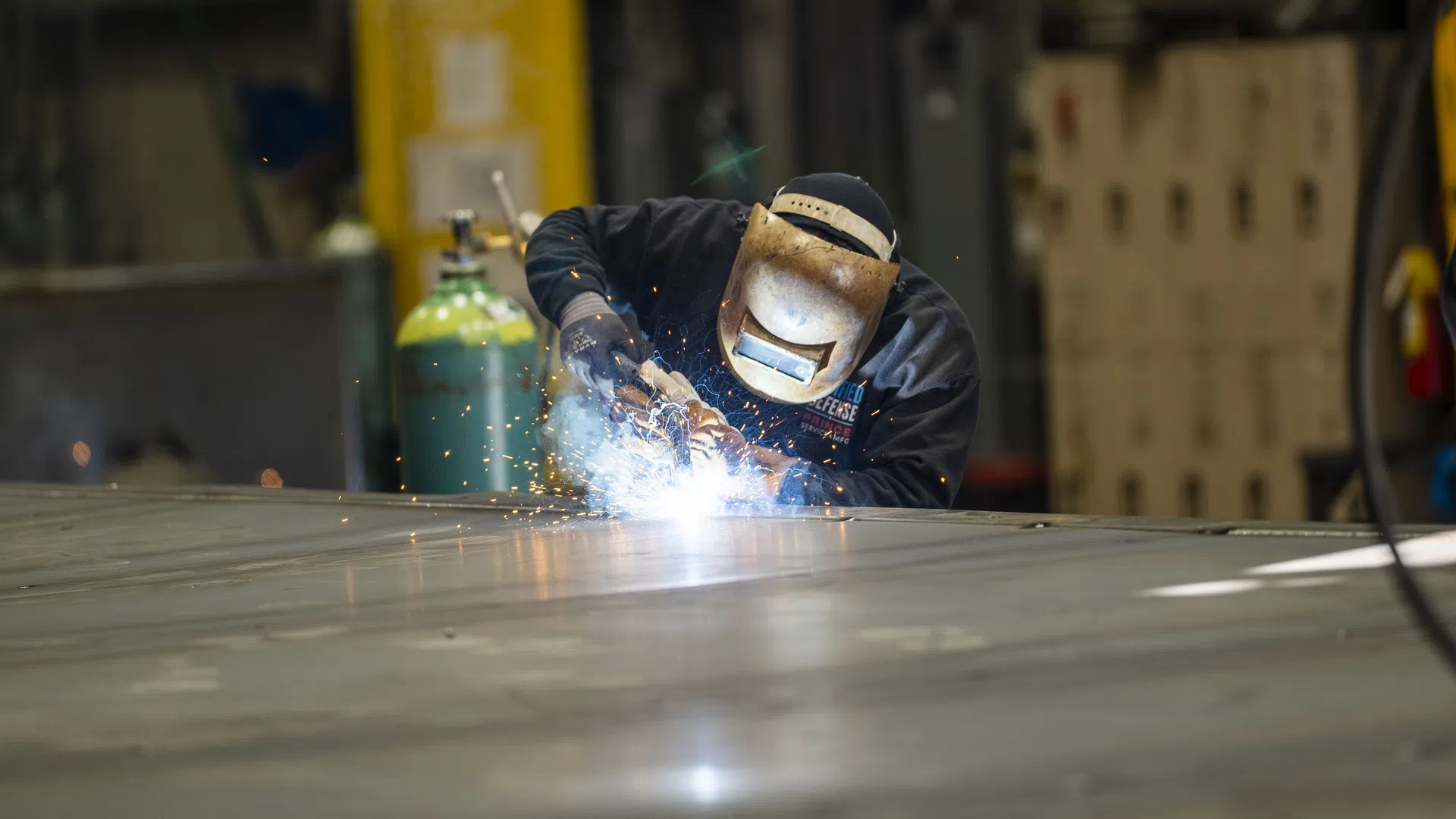 Welder wearing protective gear sparks while welding a large metal surface in an industrial workshop.