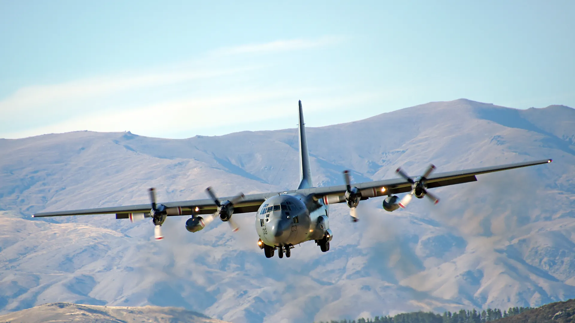 Military transport aircraft flying low over mountainous terrain with blue sky background