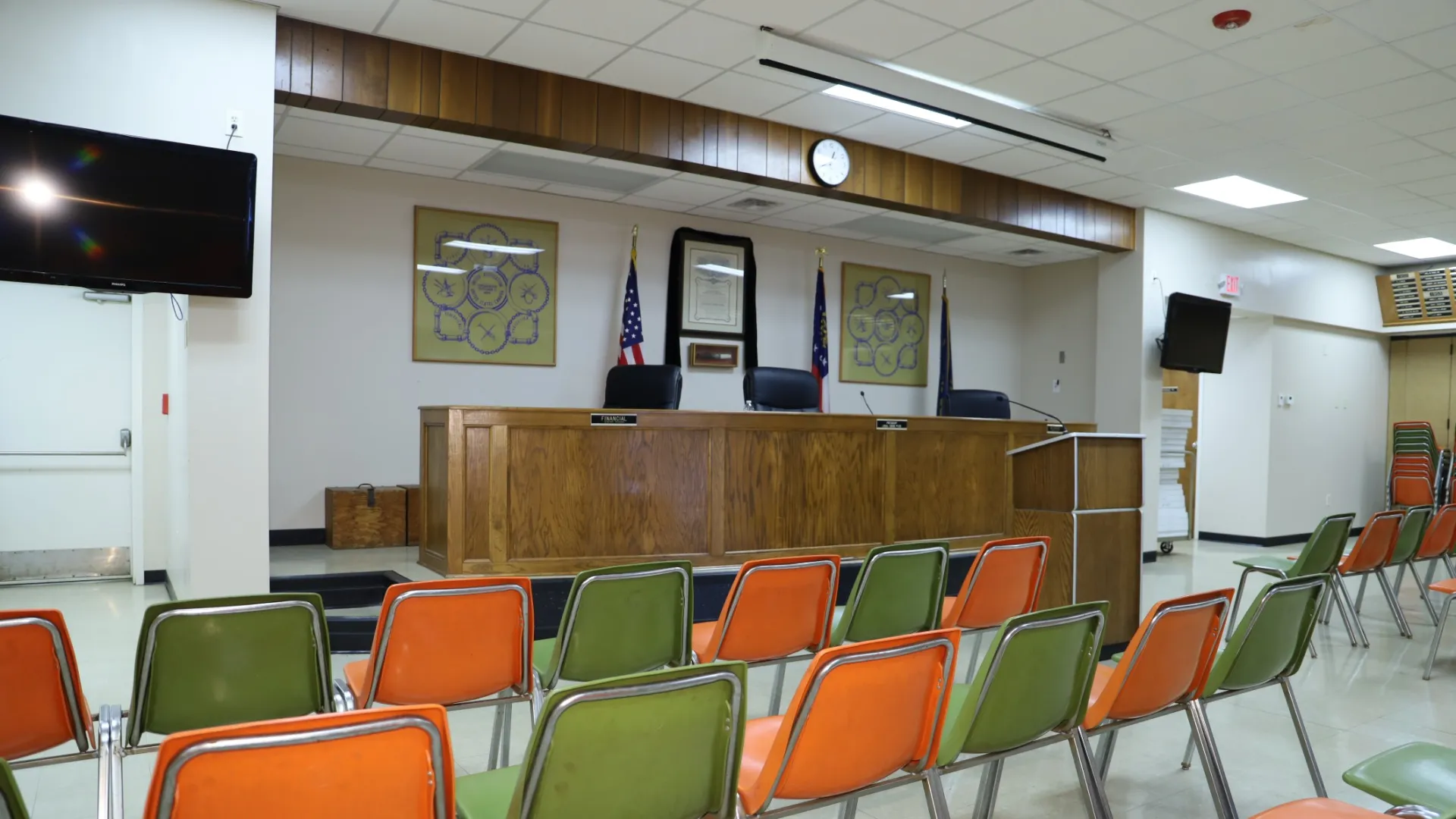 Interior of an empty meeting room featuring chairs and a wooden panel behind the dais.