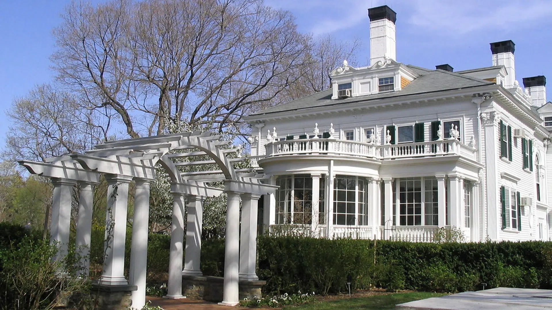 Historic white colonial mansion with pergola entrance and lush green garden under blue sky.