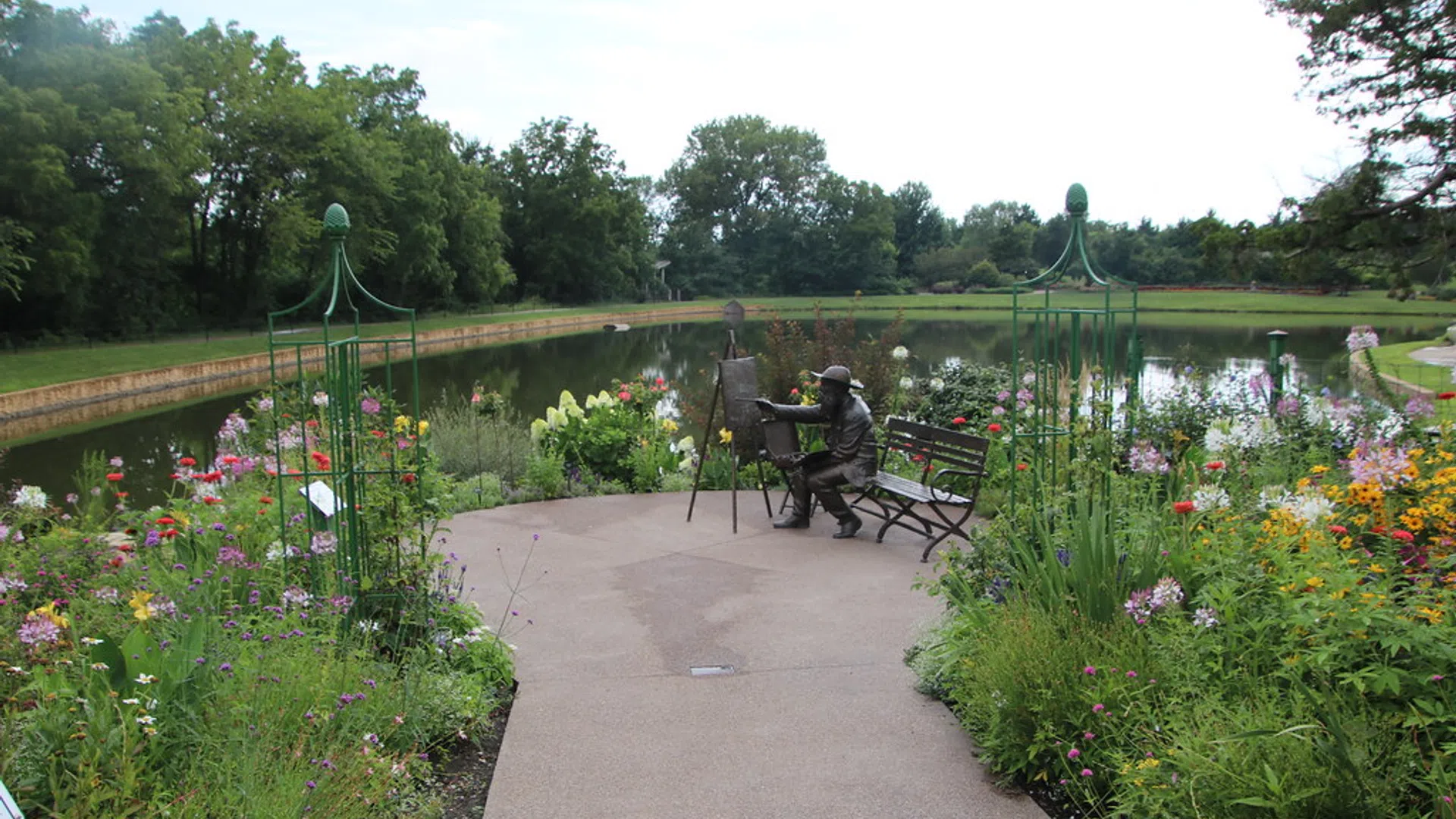 Garden path leading to a pond with a bronze statue of a painter at an easel surrounded by colorful flowers.