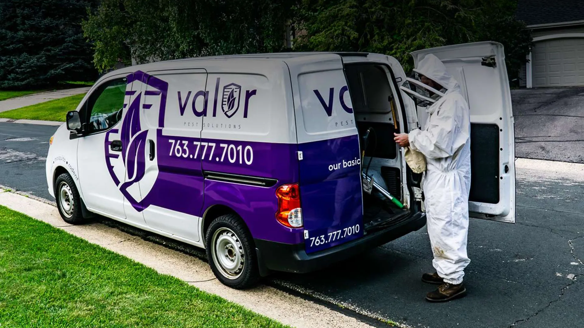 Pest control technician in protective suit loading equipment into Valor Pest Solutions service van in suburban neighborhood