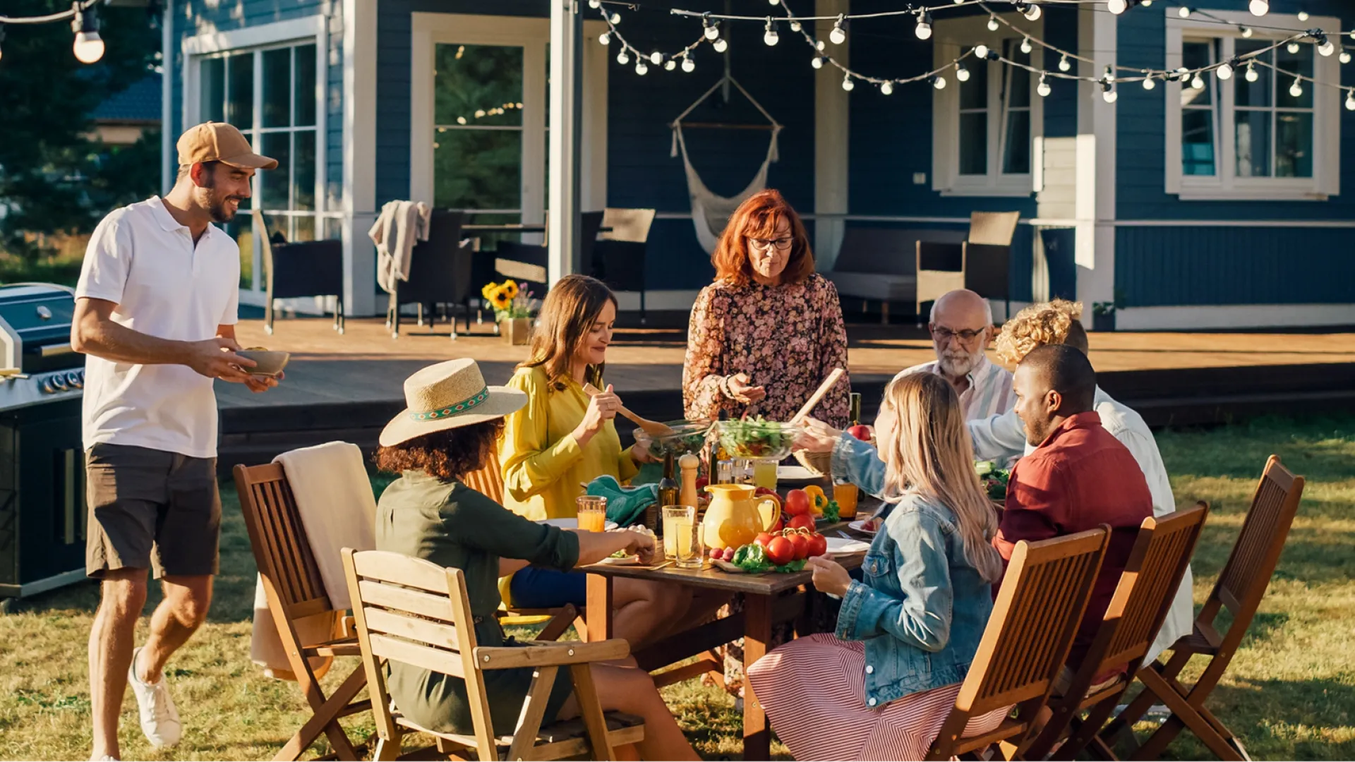 Group of people enjoying an outdoor meal together around a wooden table with string lights in backyard