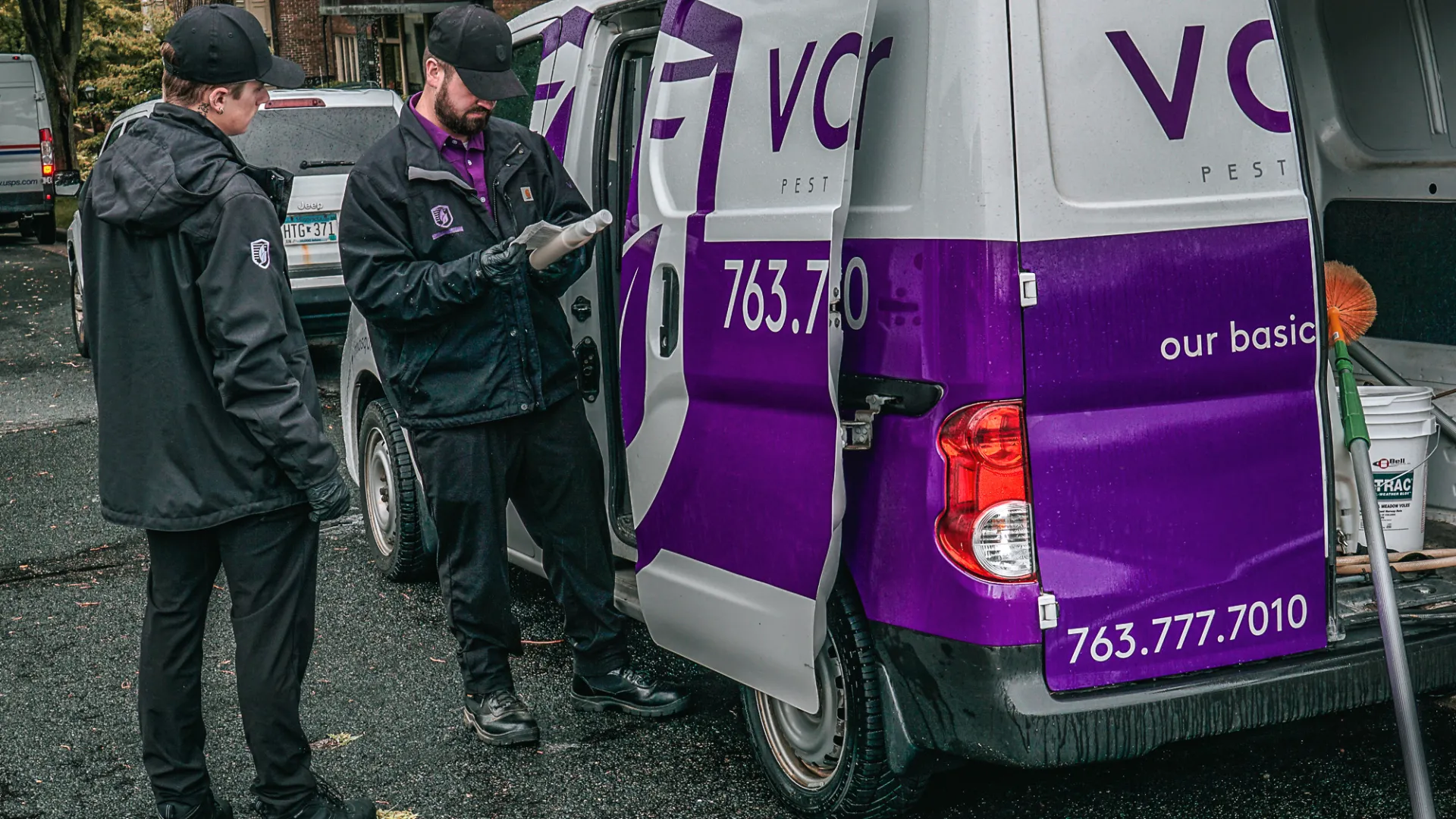 Two pest control workers in black uniforms standing near a purple and white pest control van on a residential street.