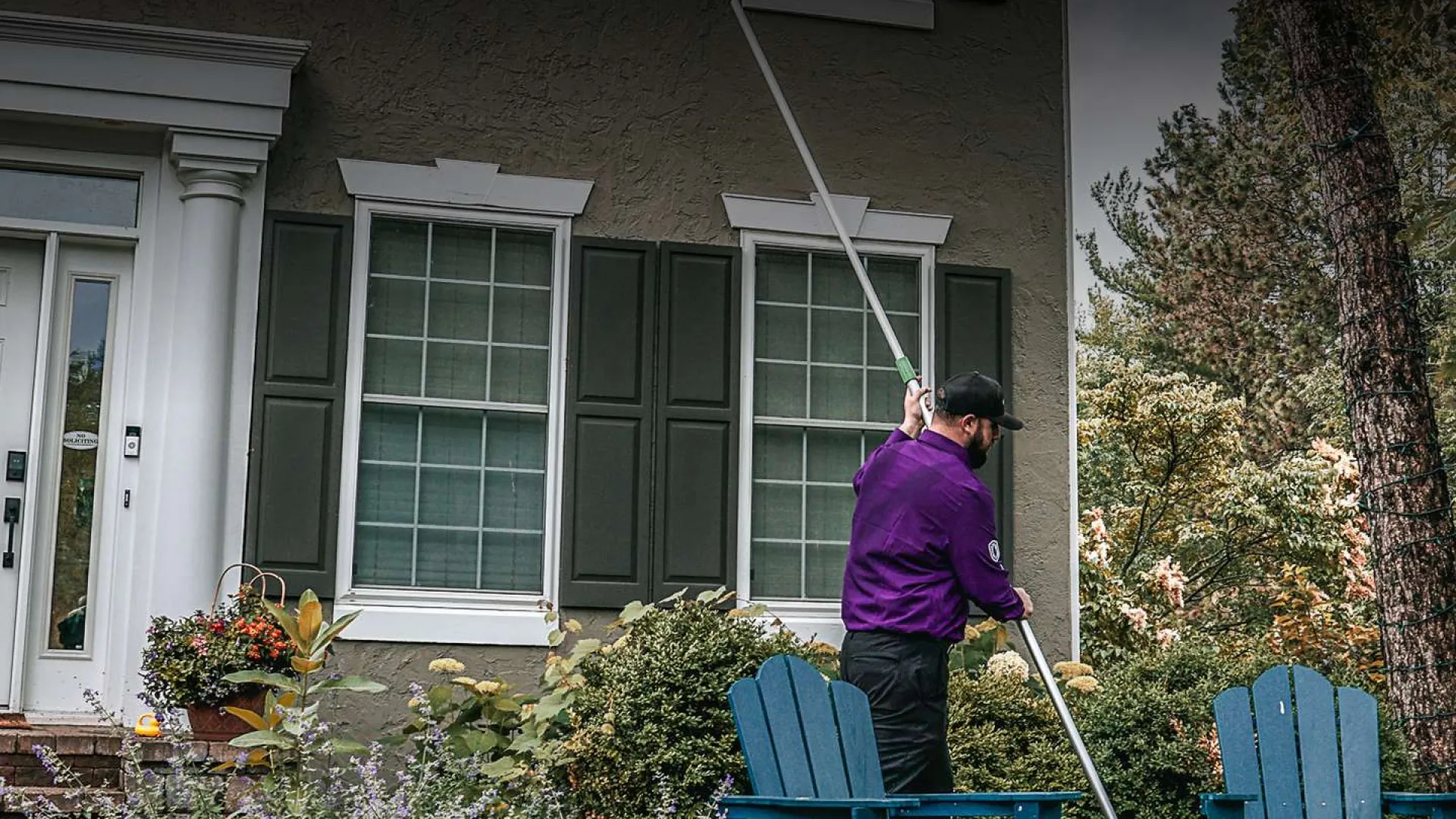 Man in purple shirt using a long pole to clean exterior windows of a suburban house with garden and chairs.