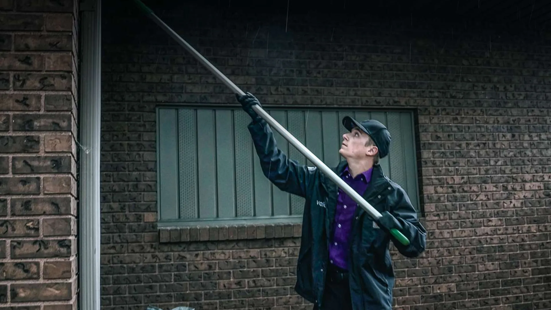 Worker wearing uniform and cap uses a long pole to clean or inspect the exterior of a brick building on a rainy day.