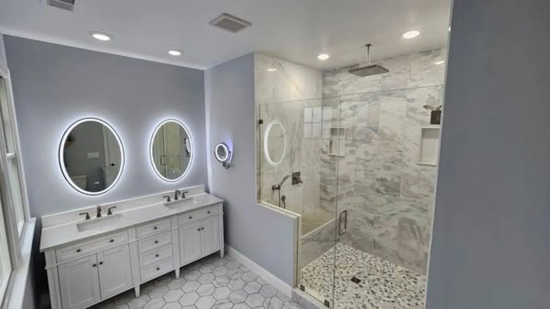 Modern bathroom with double white vanity, illuminated round mirrors, and glass-enclosed rain shower with marble accents