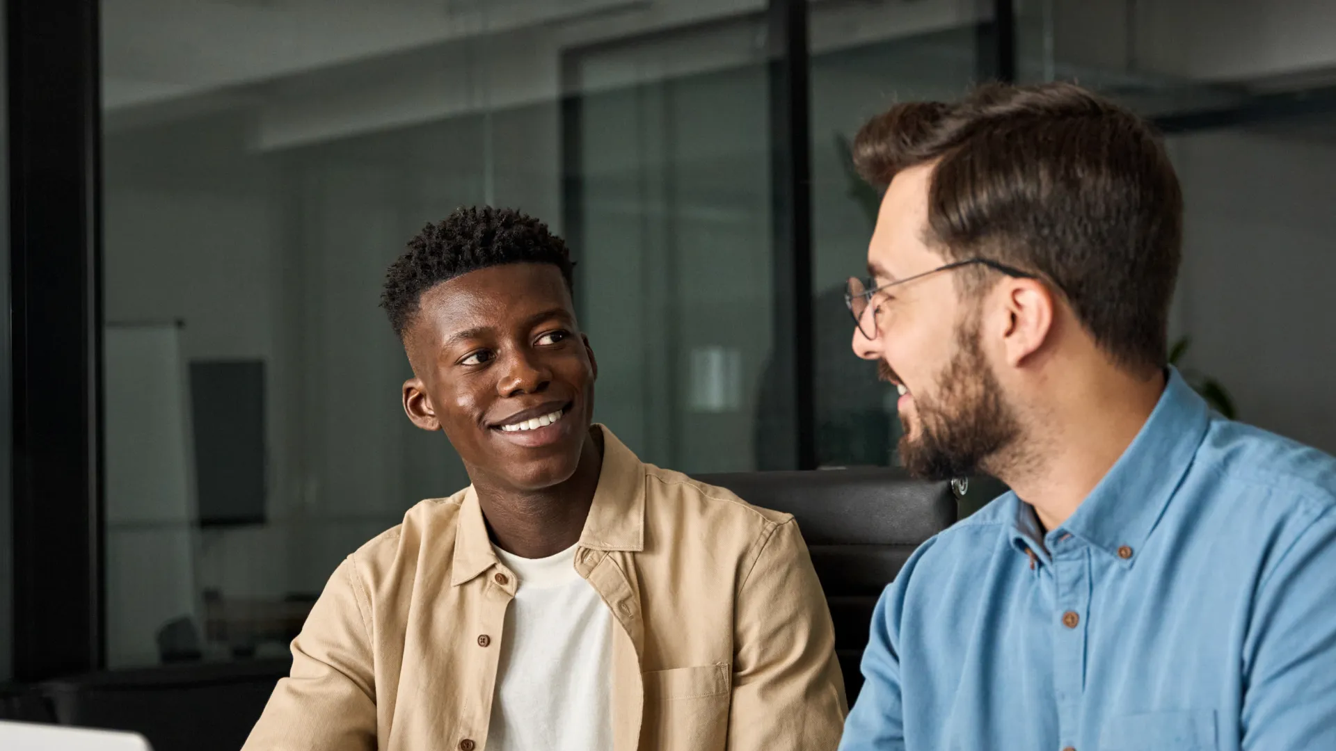 Two men in an office discussing documents with a laptop on a wooden table, smiling and engaged in conversation