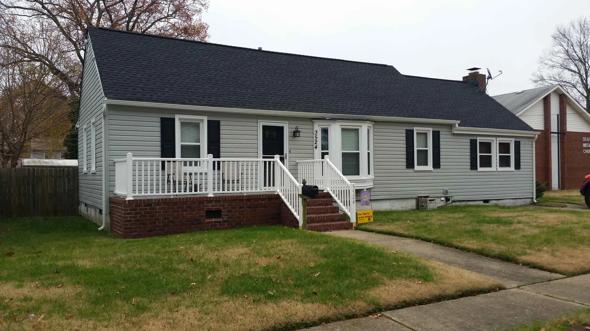 Single-story gray house with a black roof, white railing porch, and a brick foundation on a grassy lawn.