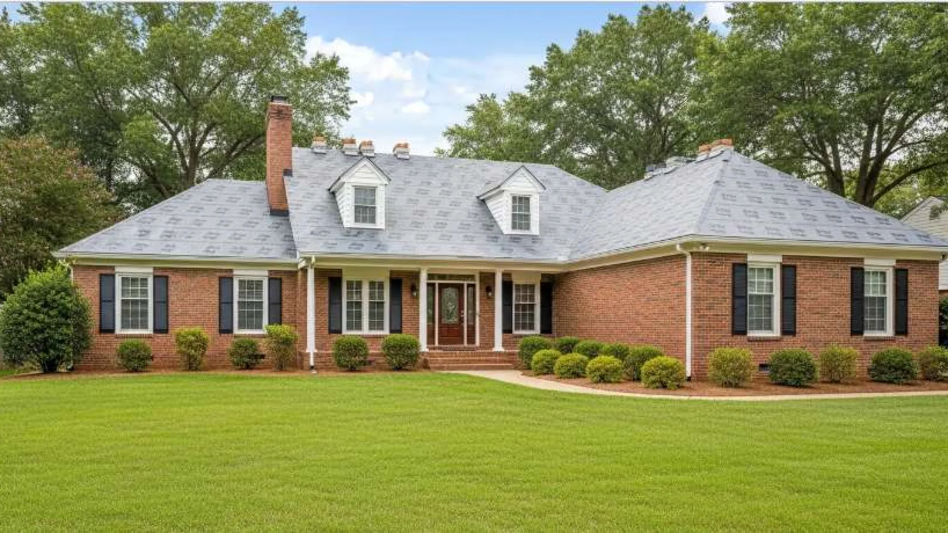 Brick single-story house with gray roof, white columns, black shutters, and green lawn in front under blue sky.