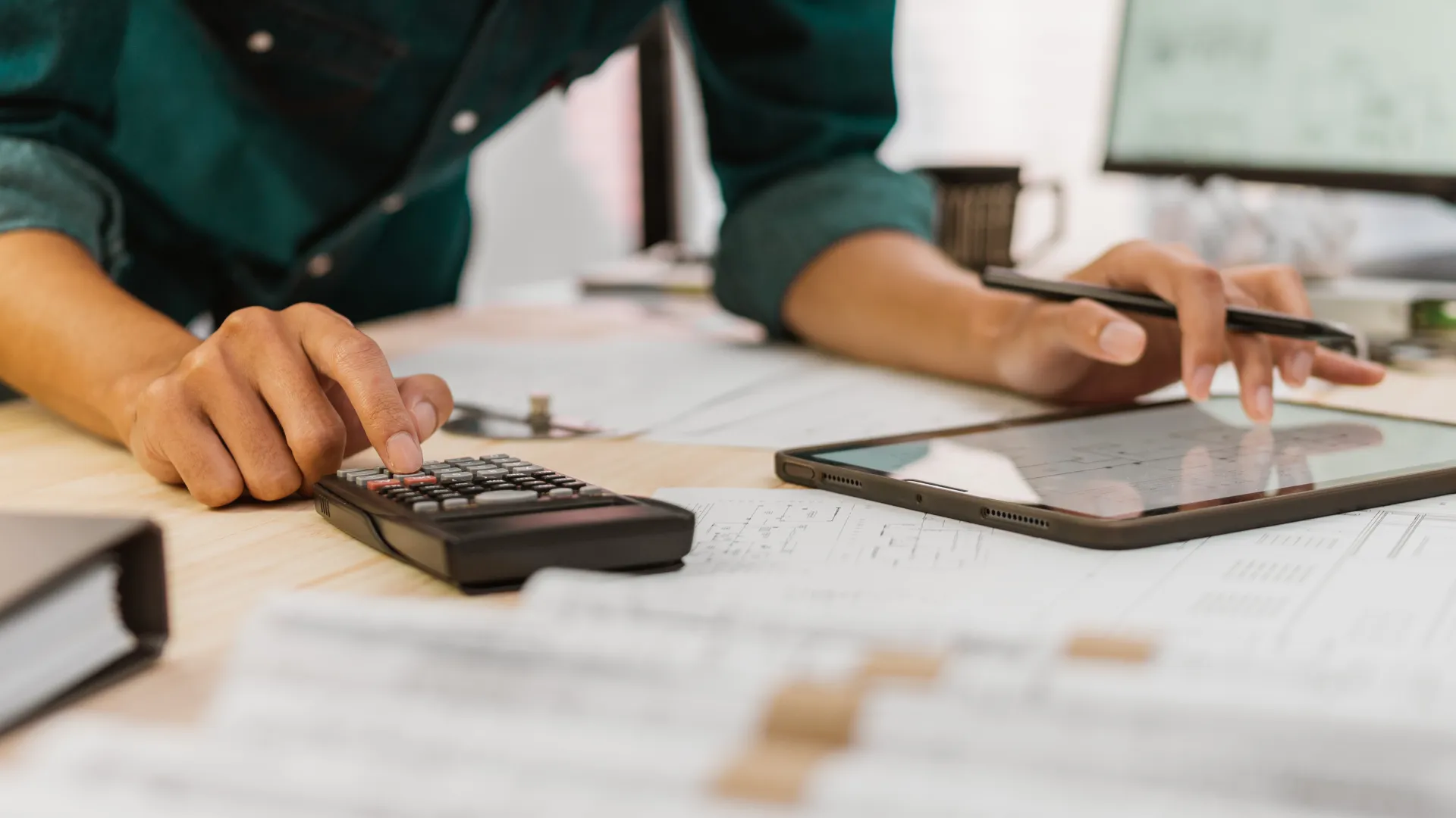 Person using calculator and tablet with pen over documents on a wooden desk for work or study.