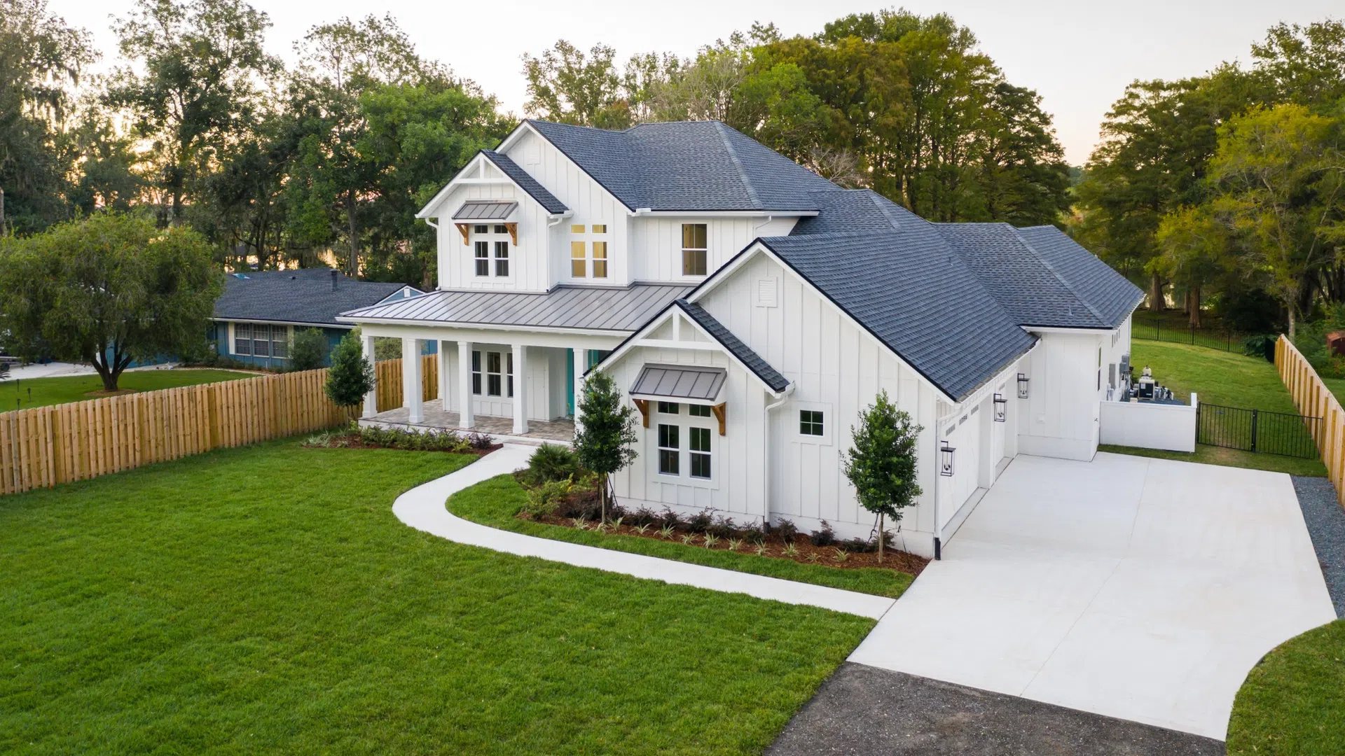 Large modern white farmhouse with gray roof, spacious porch, green lawn, and surrounding trees at dusk.