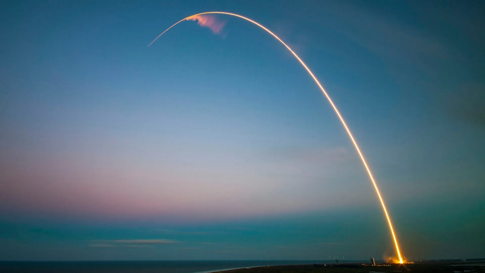 Long-exposure photo of a rocket launch creating a bright arc over a coastal landscape at dusk.