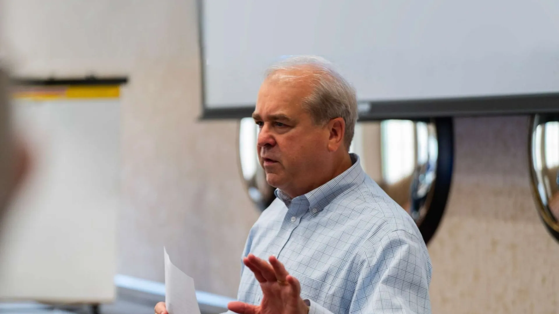 Man in checkered shirt holding a paper and speaking in a conference room with a whiteboard and screen.