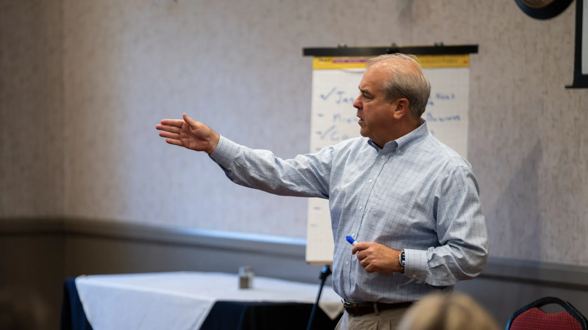 Mature man in button-up shirt speaking and gesturing during a meeting with flipchart and seated audience.