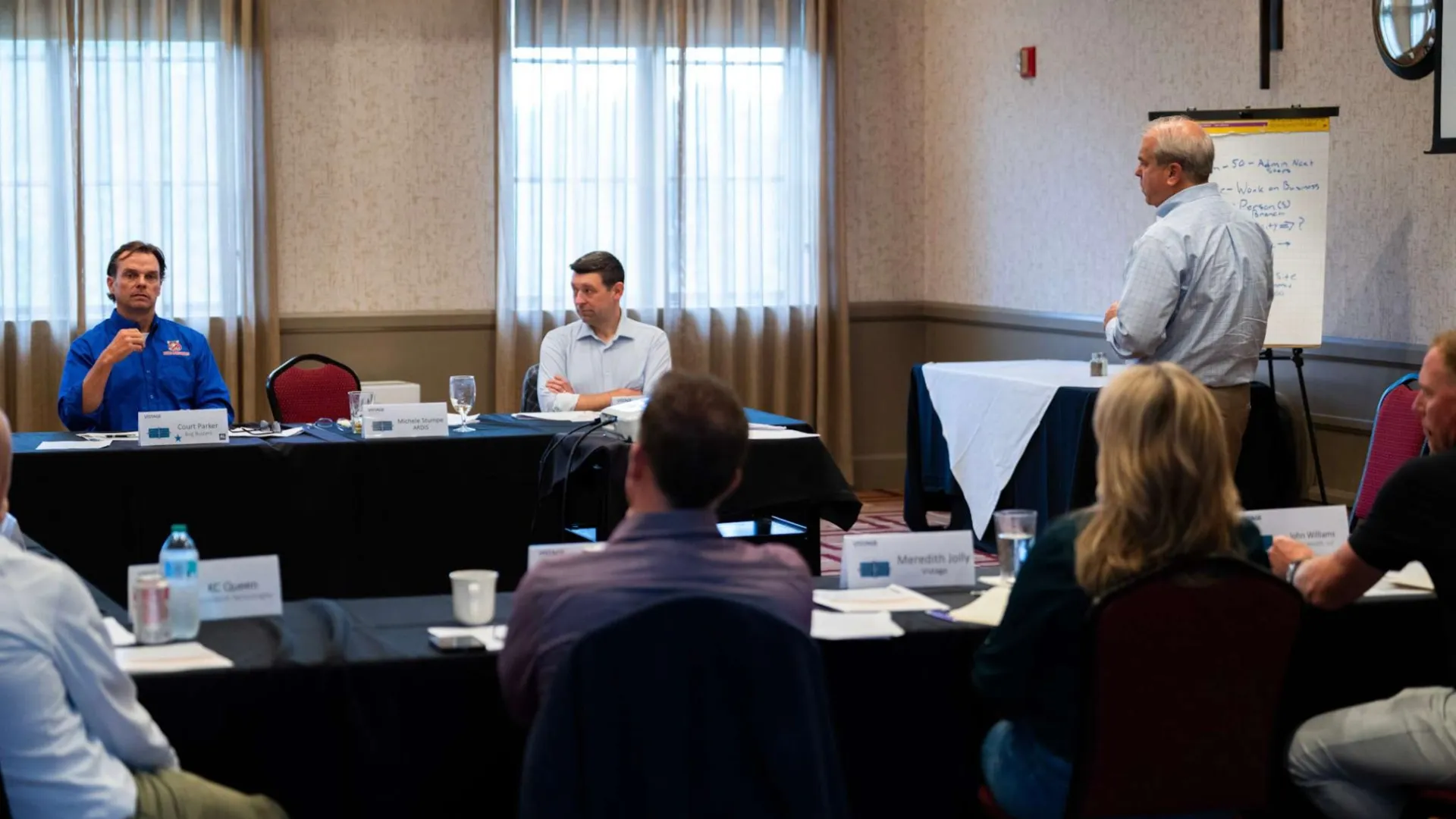 Business meeting with participants seated around tables, one person standing near flip chart speaking.