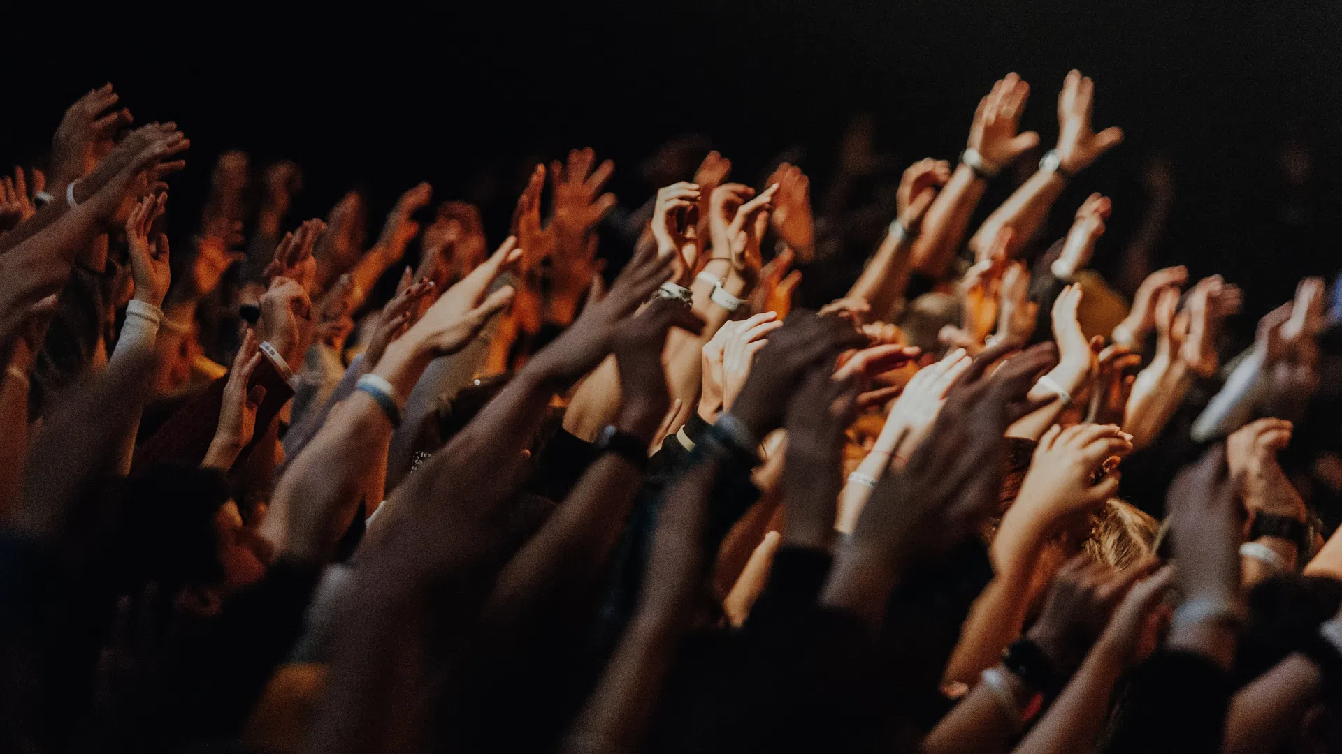 Crowd of people raising hands enthusiastically at a concert or event with dark background and warm lighting.