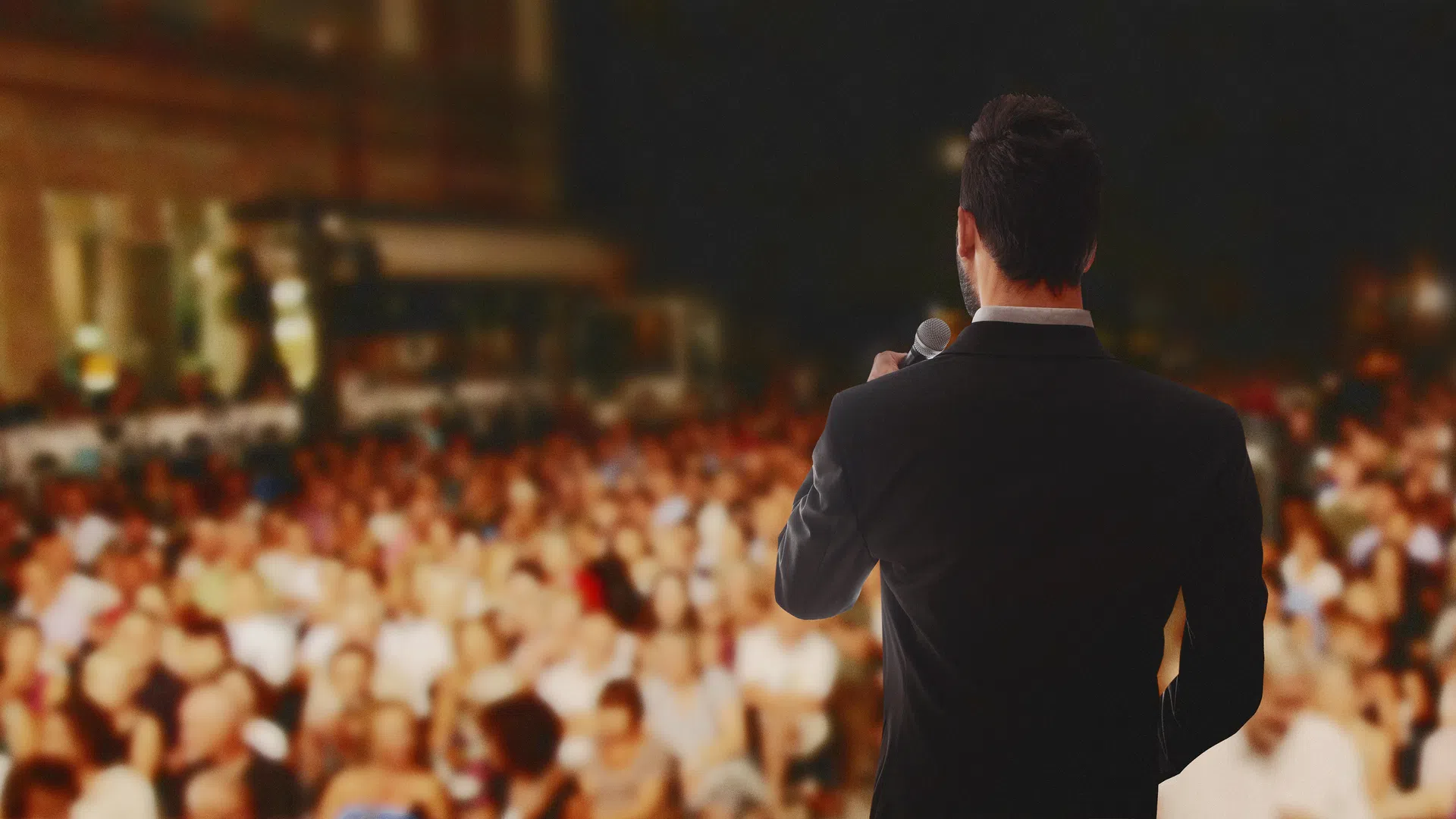 Speaker in a suit addressing a large crowd at an outdoor event during the evening.