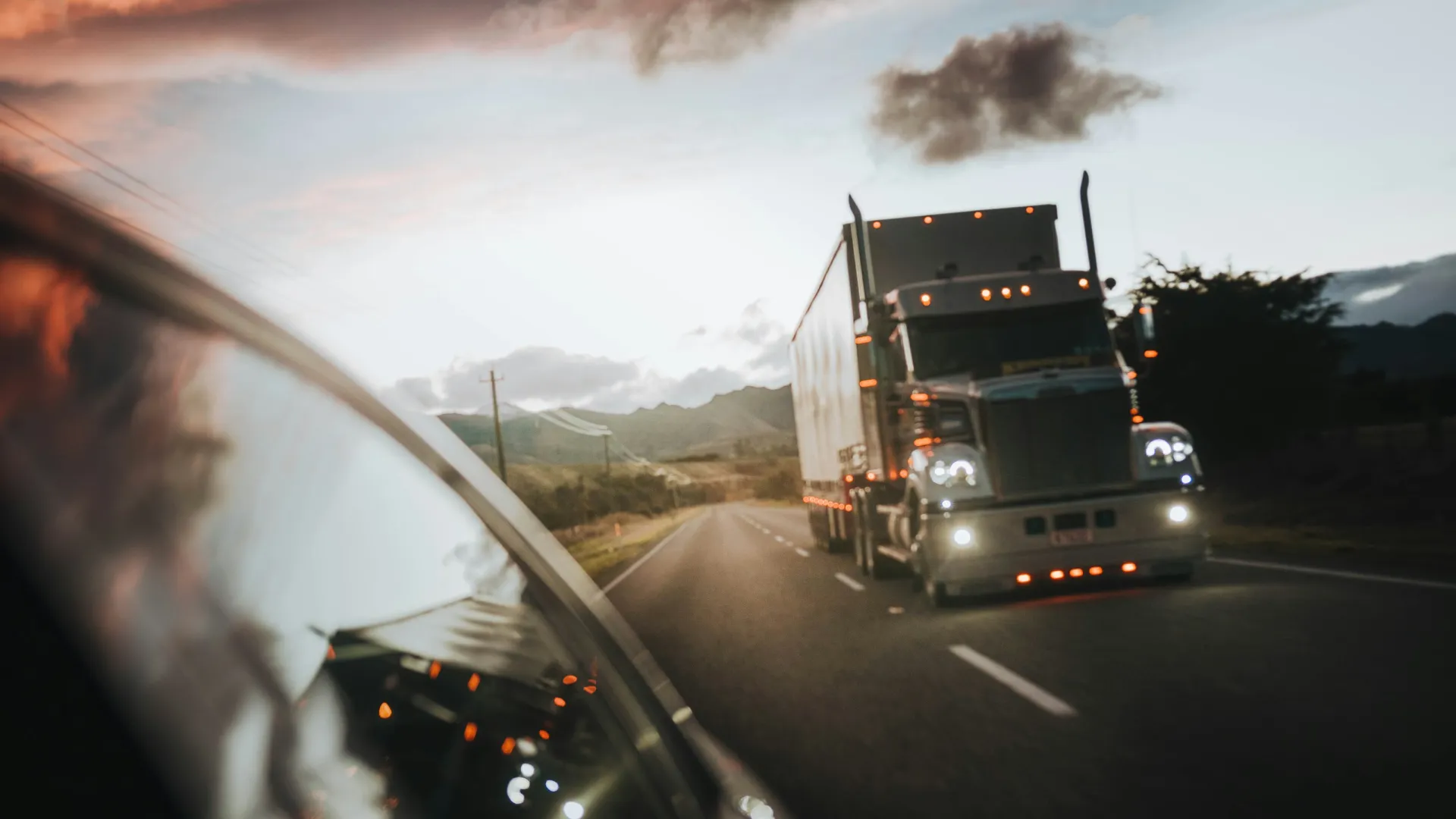 Semi-truck driving on an open highway at sunset with mountains and clouds in the background