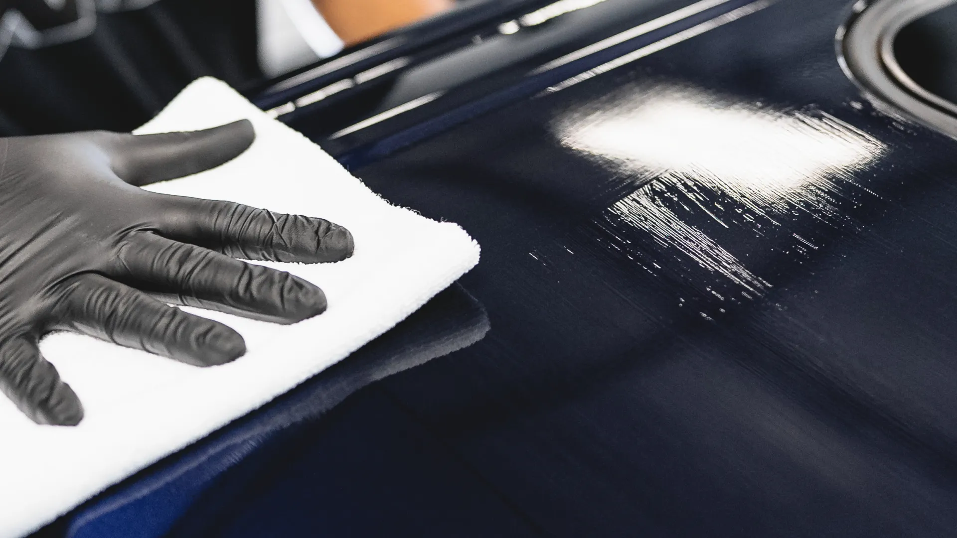 Person wearing black gloves polishing a shiny dark blue car hood with a white cloth indoors.