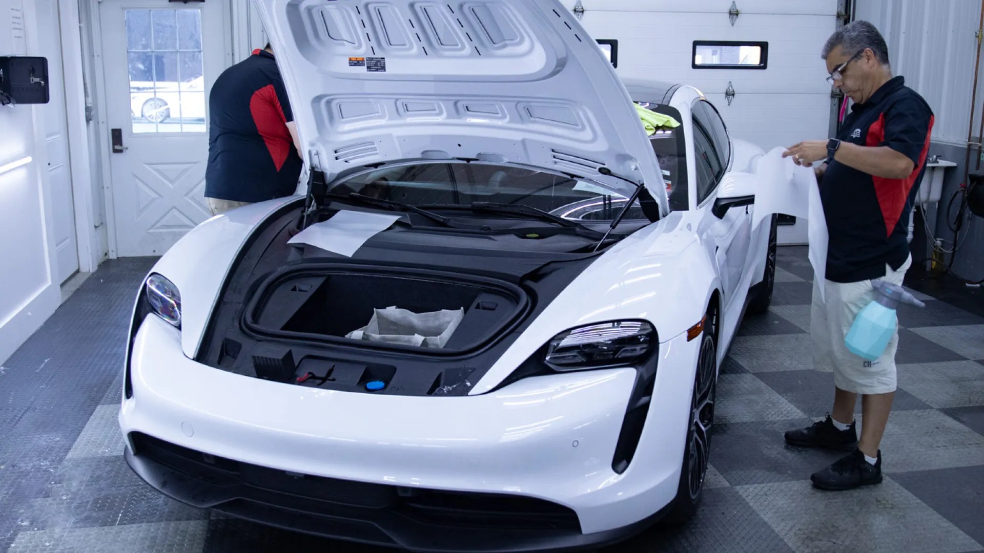 White sports car with open front trunk being inspected by two men inside a garage with checkered floor.