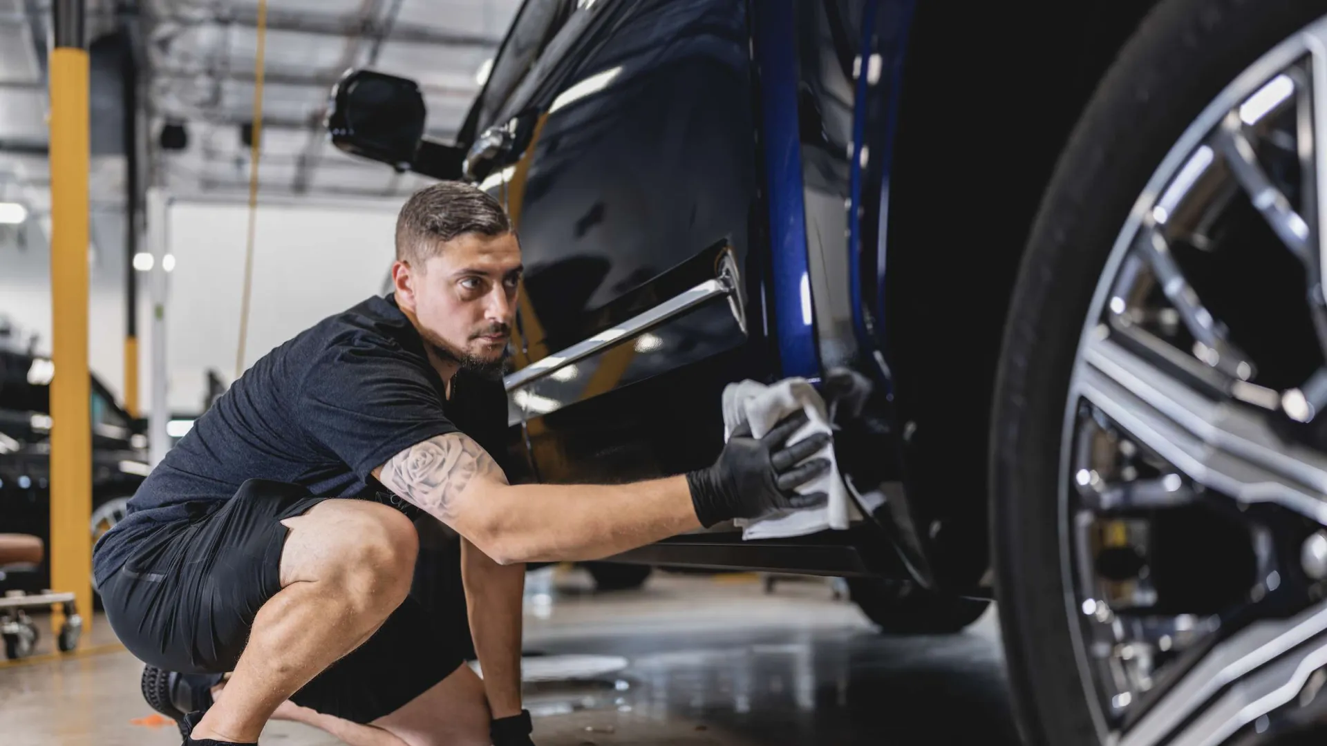 Man wearing black gloves carefully polishing a dark blue car in a well-lit garage.